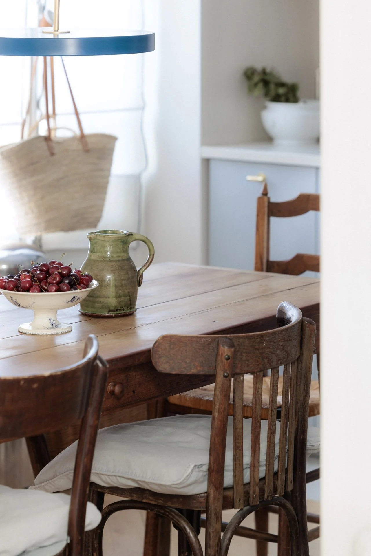 Table à manger en bois ancien avec coupe de cerises, pichet en céramique verte et chaises vintage, suspension bleue et cuisine en arrière-plan