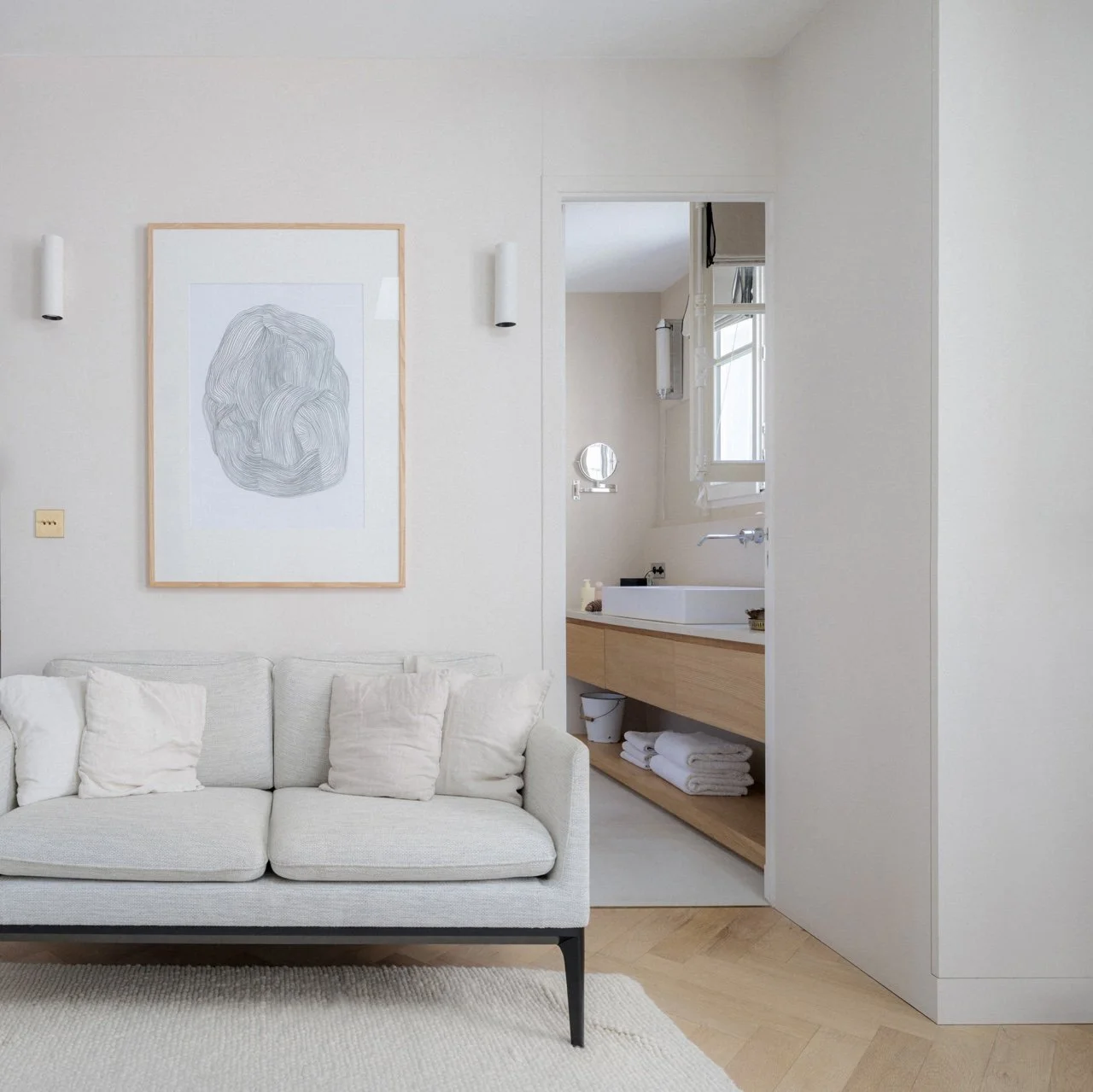 Salon minimaliste avec canapé beige, coussins et illustration abstraite encadrée, vue sur salle de bain avec meuble vasque en bois clair