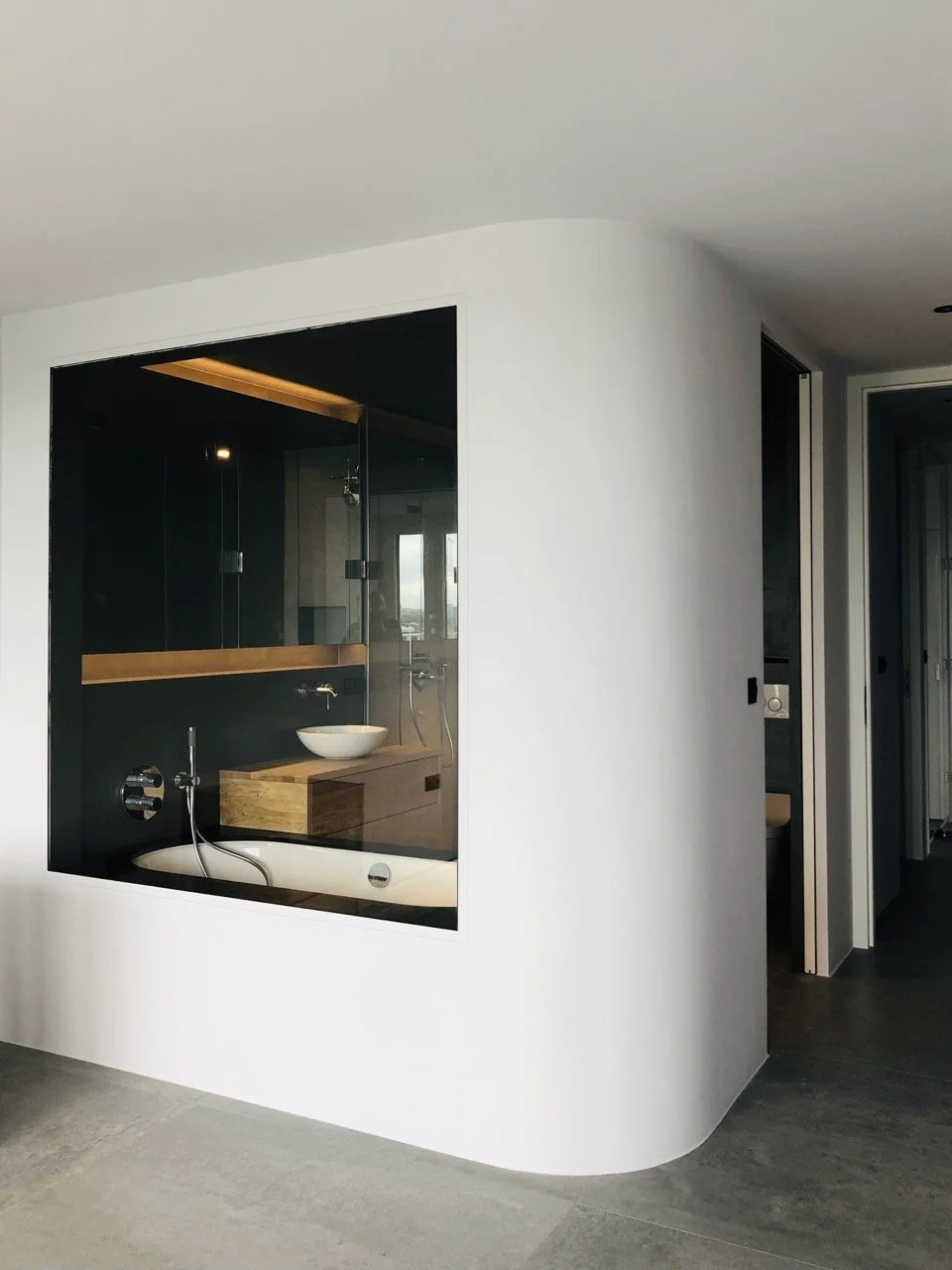 Angle arrondi et grande vitre ouvrant sur la salle de bain de la chambre principale, meuble vasque en bois et sol en béton ciré