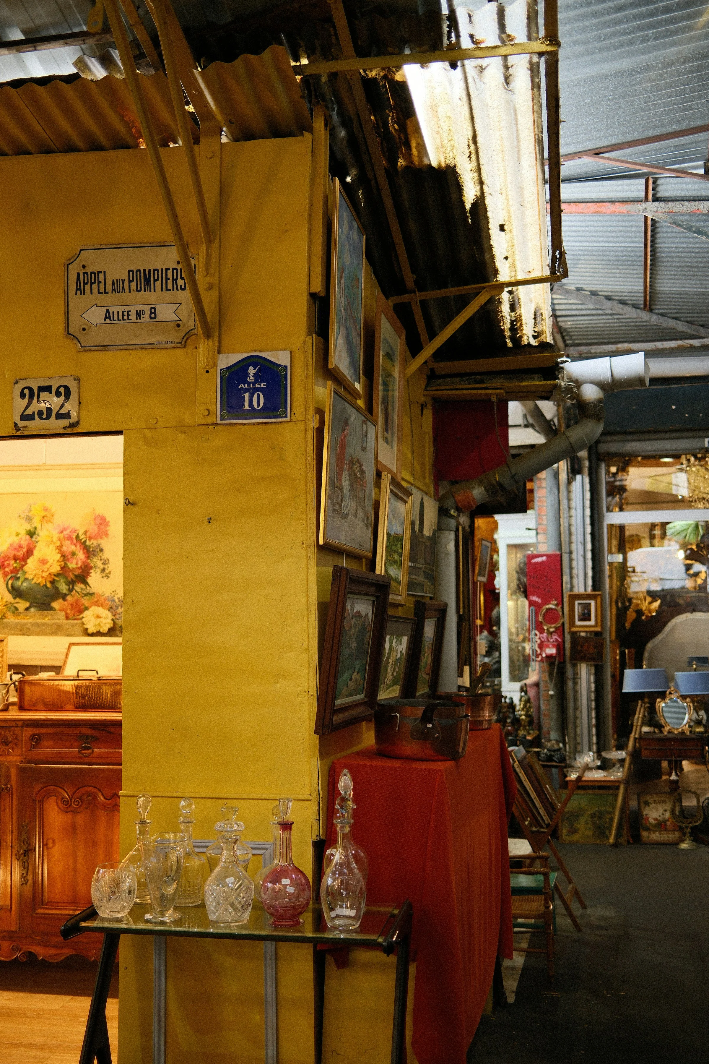 Étal d'antiquaire aux Puces de Saint-Ouen : murs jaunes, tableaux anciens et carafes en verre