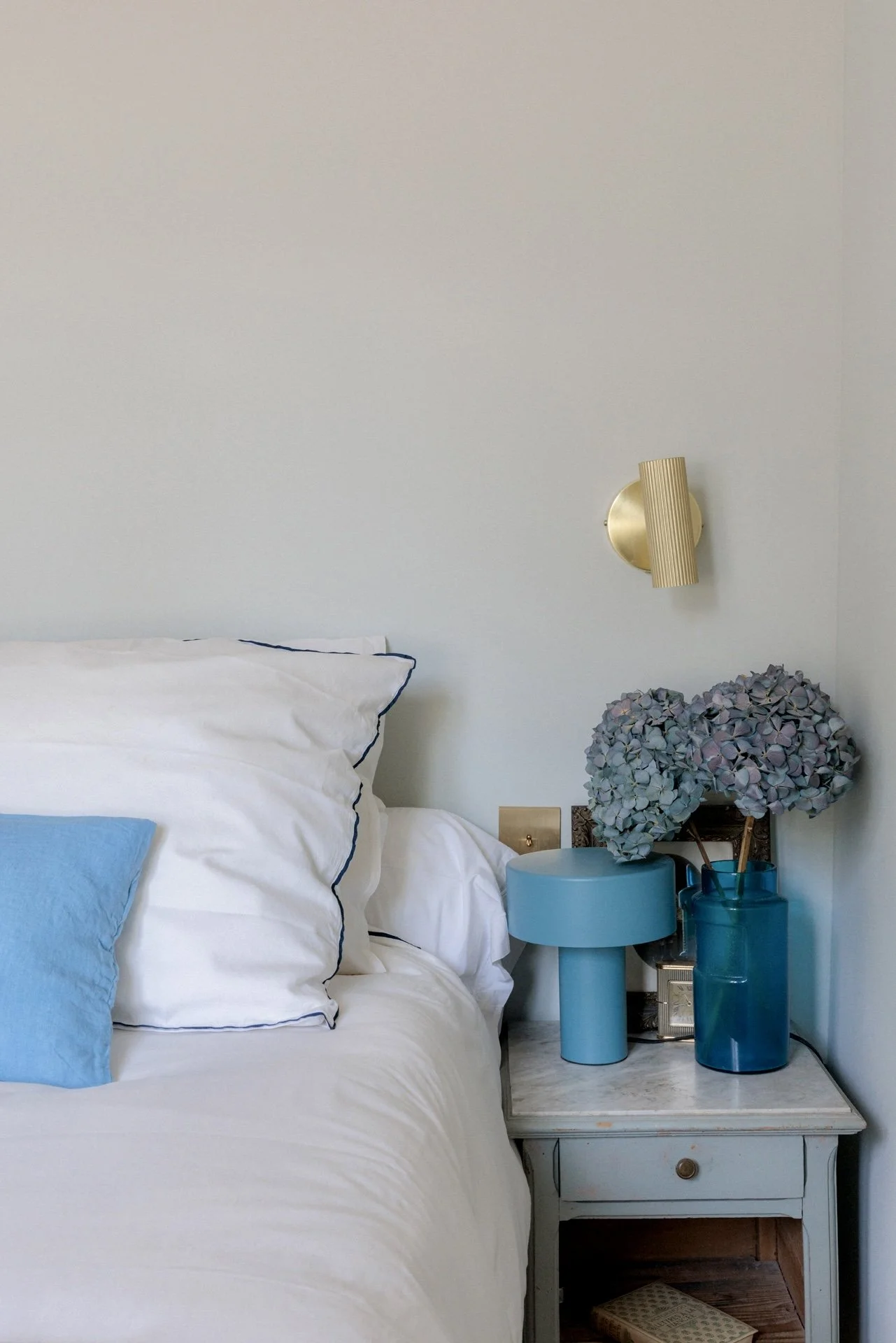 Table de chevet vintage avec lampe bleue, vase en verre et hortensias séchés, à côté du lit au linge blanc à liséré marine