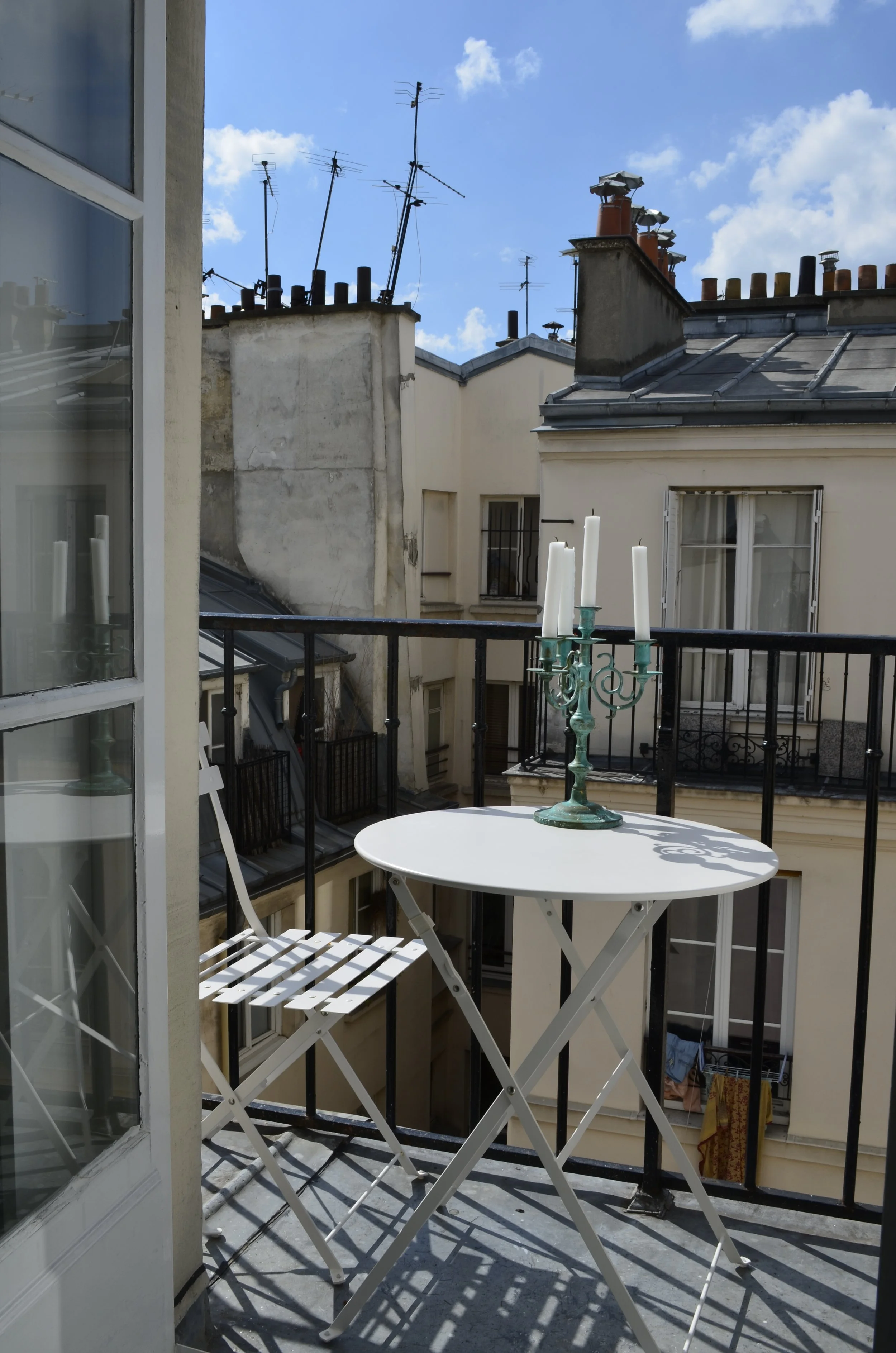 Balcon avec table bistrot ronde et chaises pliantes, garde-corps en fer forgé et vue sur les toits de Paris depuis le sixième étage