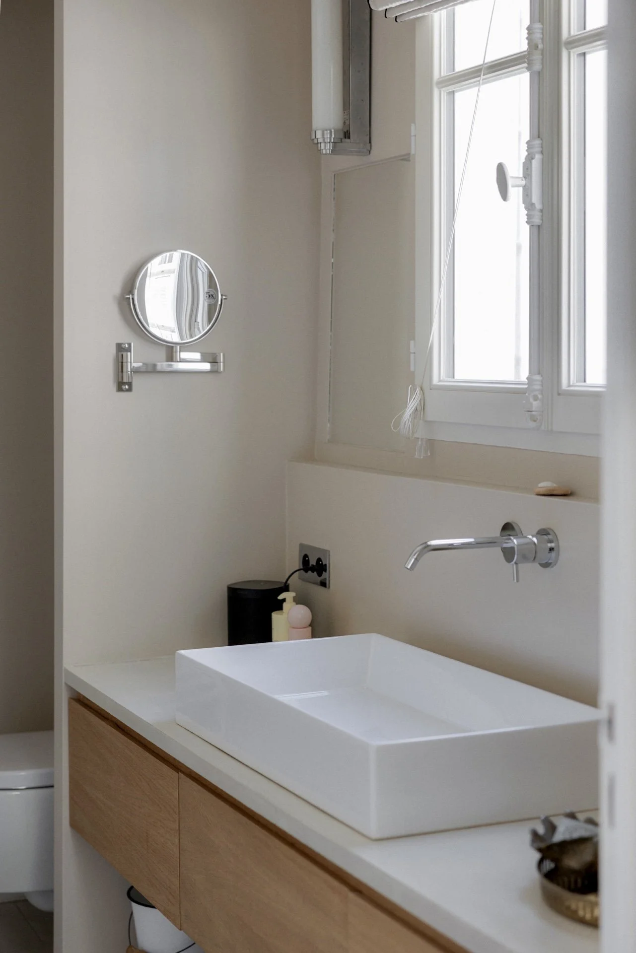 Salle de bain avec vasque rectangulaire blanche à poser, robinetterie murale chromée, miroir grossissant et meuble vasque en chêne et béton ciré devant la fenêtre