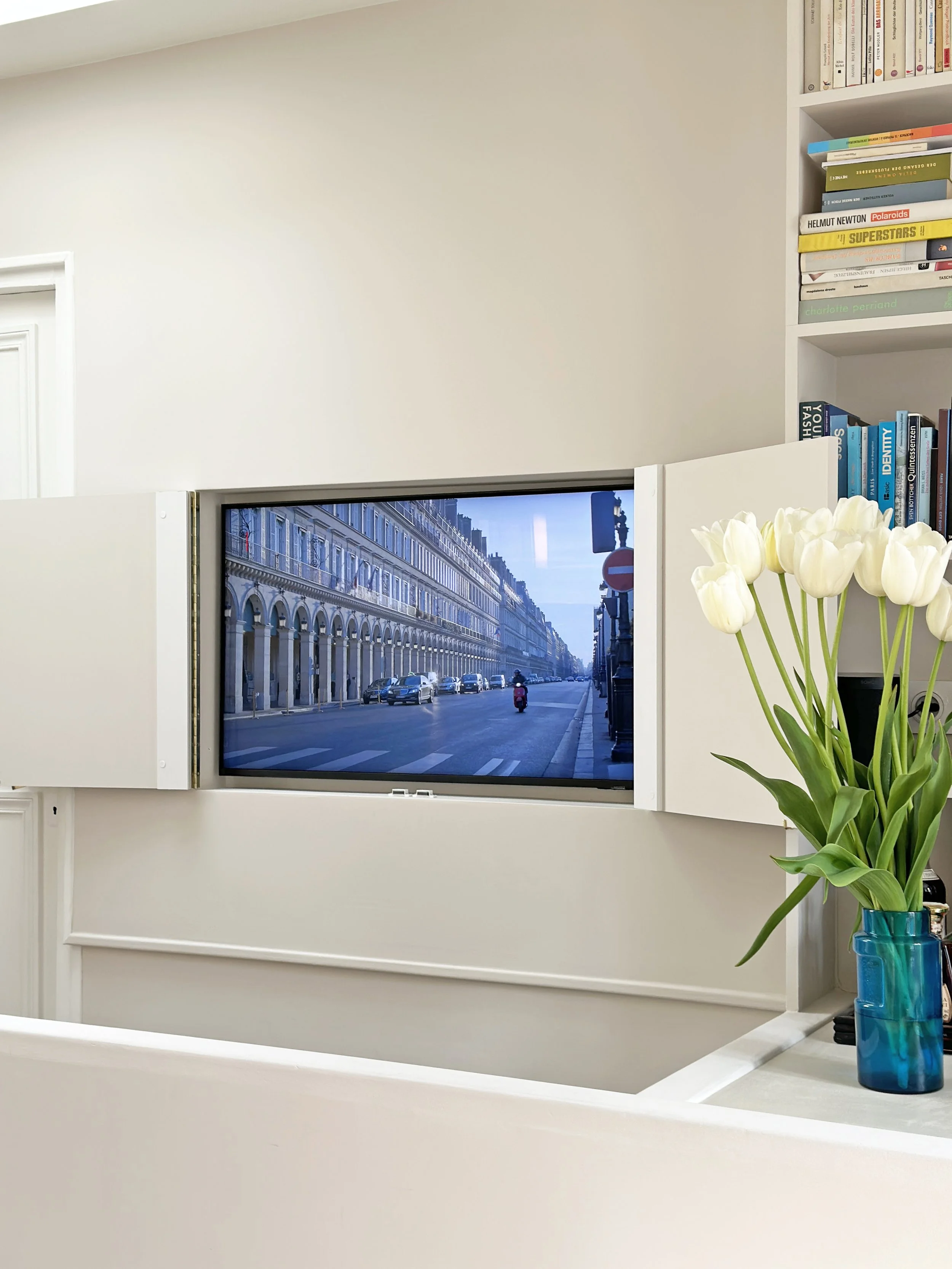 Télévision escamotable sortant d'un meuble bas laqué blanc devant la bibliothèque, bouquet de tulipes blanches