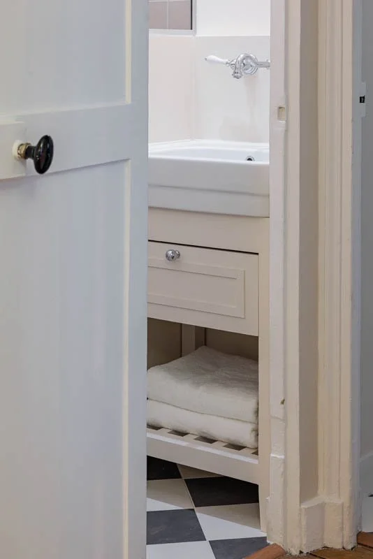 Vue depuis la porte sur une salle de bain épurée, vasque et robinetterie chromée d'inspiration classique, meuble sous-vasque laqué crème avec claustra bas et linge de toilette plié, sol en damier noir et blanc.