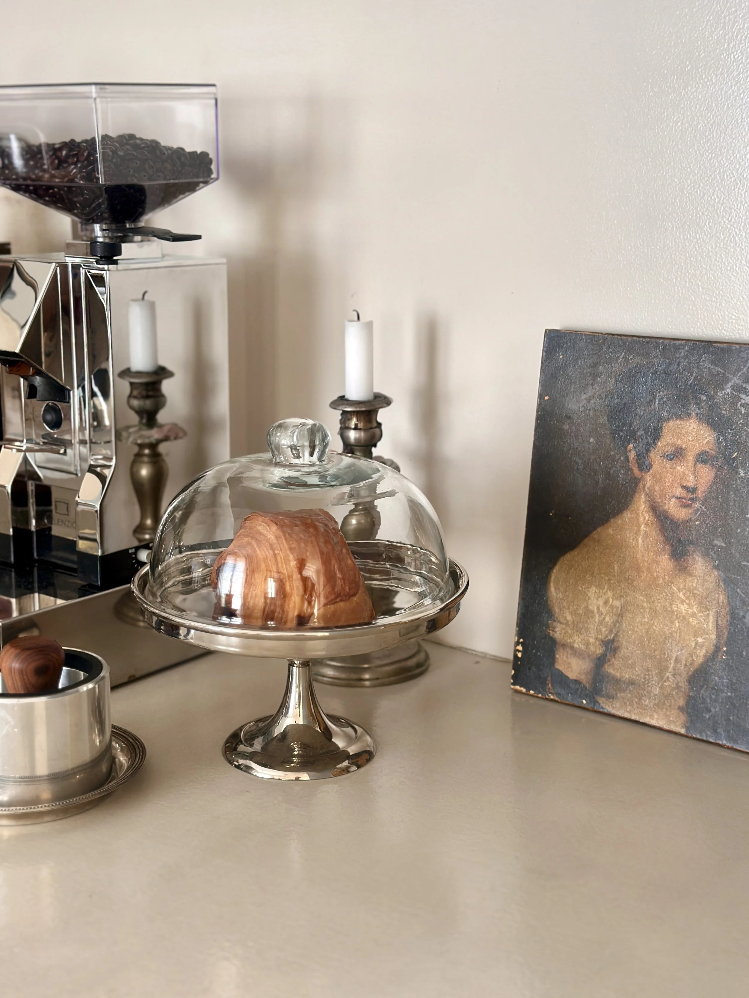 Coin cuisine avec croissant sous cloche en verre sur présentoir argenté, chandeliers, moulin à café et portrait ancien en arrière-plan
