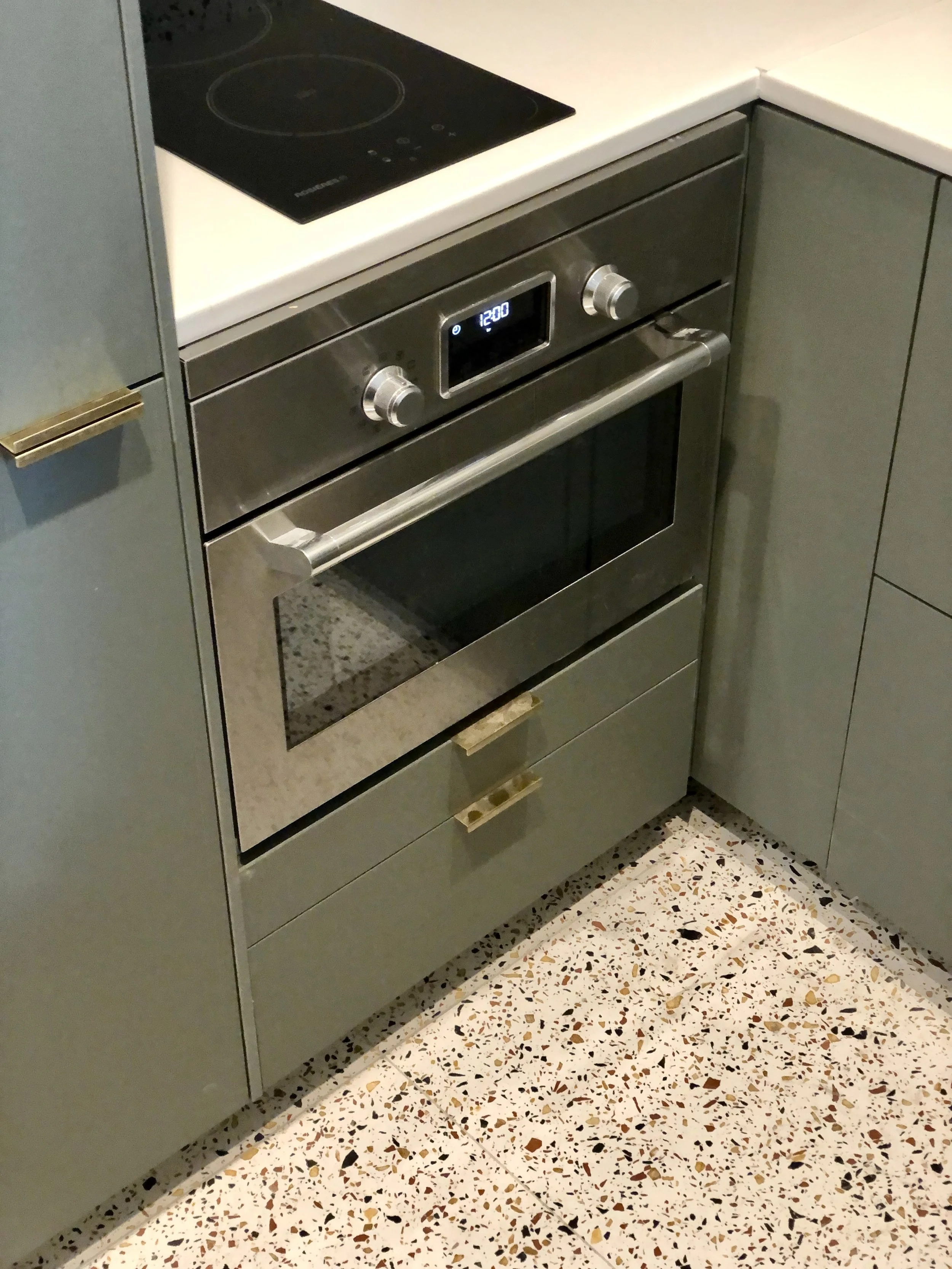 Cuisine sur mesure vert de gris avec plaque à induction, four encastré, poignées en laiton et sol en terrazzo