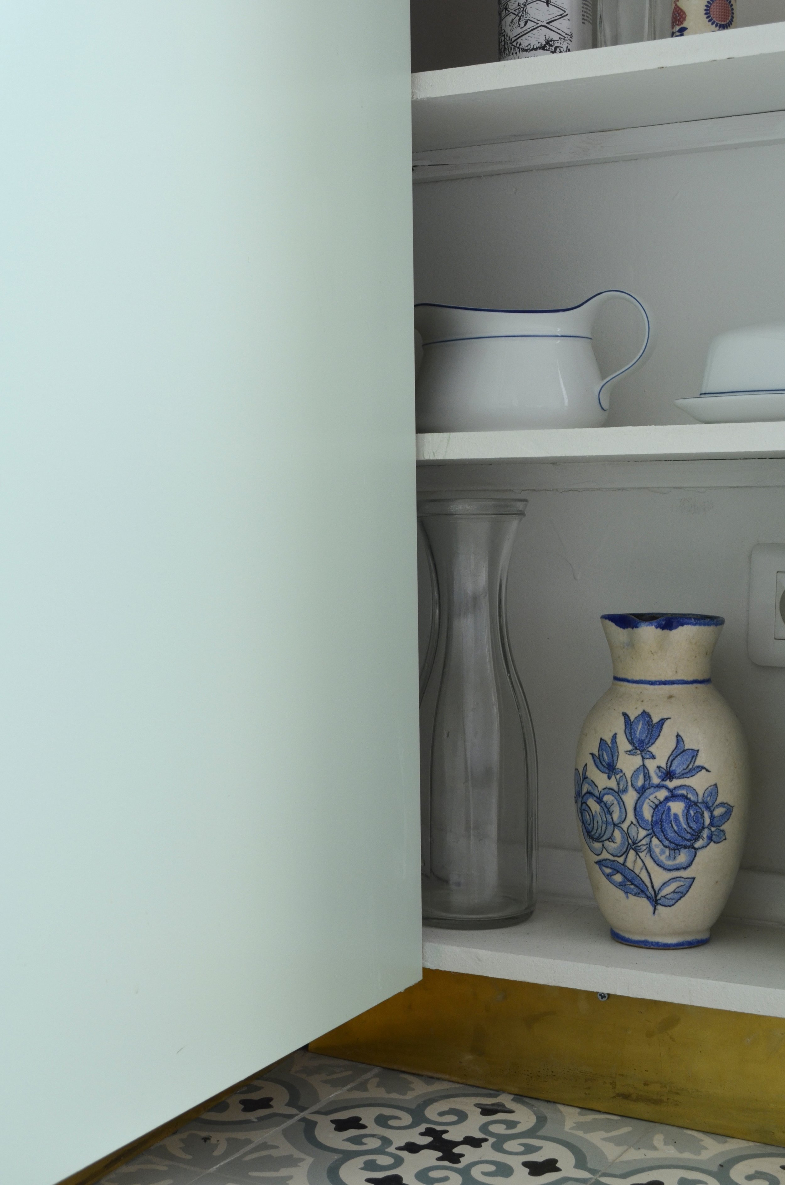 Détail d'un placard ouvert avec vaisselle, pichet blanc et vase bleu et blanc, mur vert d'eau et sol en carreaux de ciment