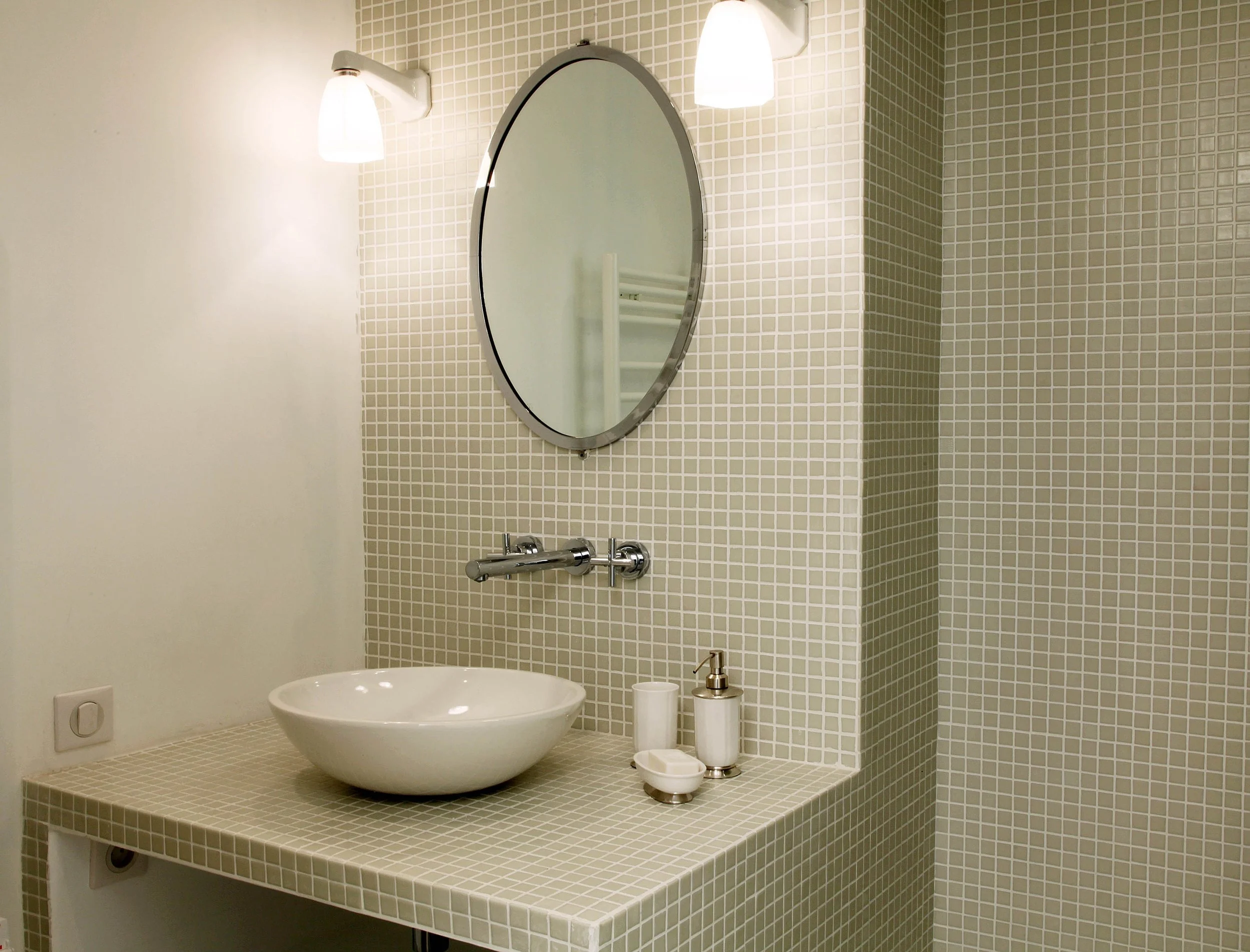 Salle de bain habillée de mosaïque avec vasque à poser blanche, robinetterie murale chromée, grand miroir ovale au cadre métal et appliques vintage