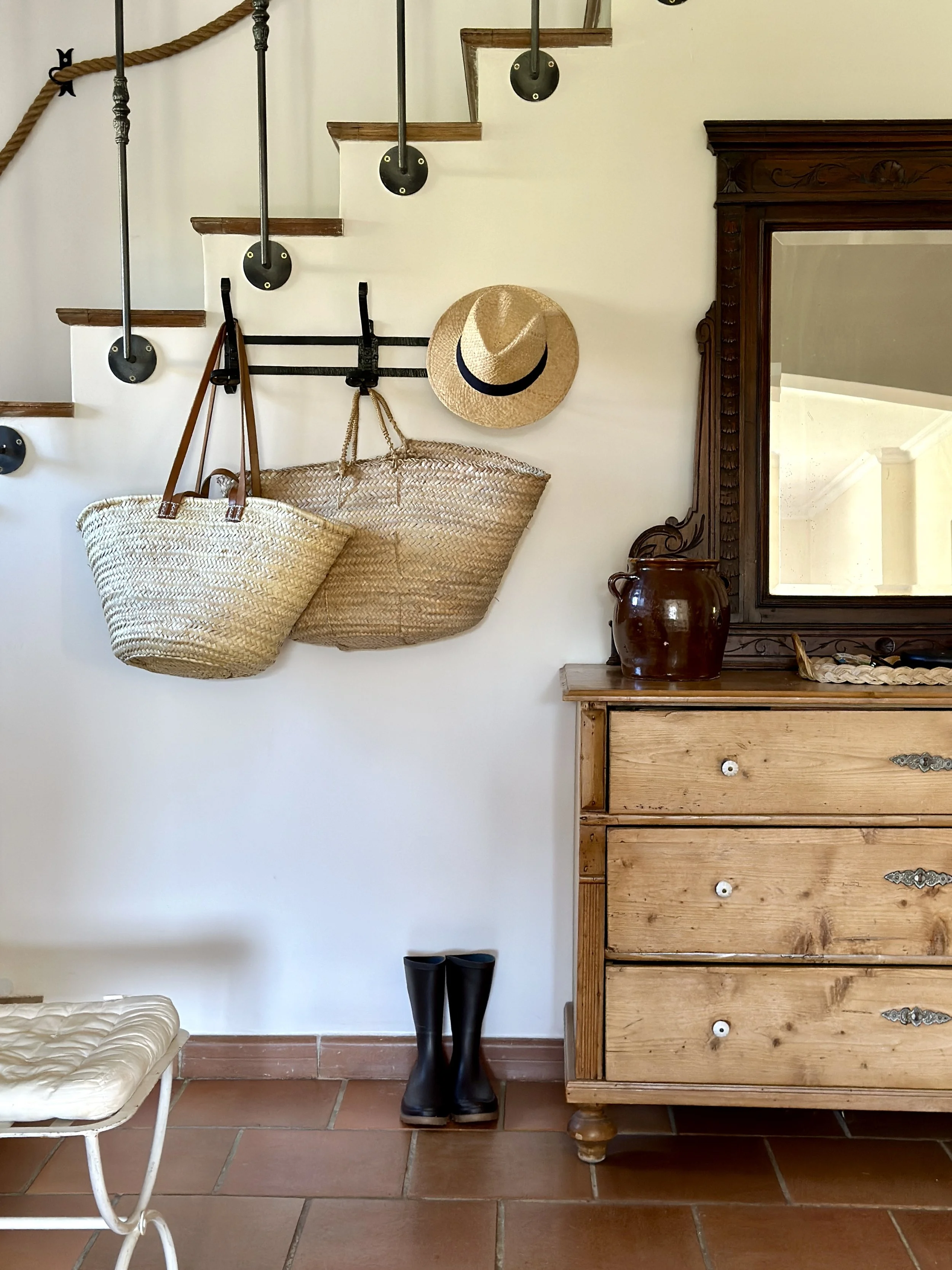 Entrée avec escalier, paniers en osier et chapeau de paille suspendus, bottes noires et commode ancienne en bois avec miroir sculpté