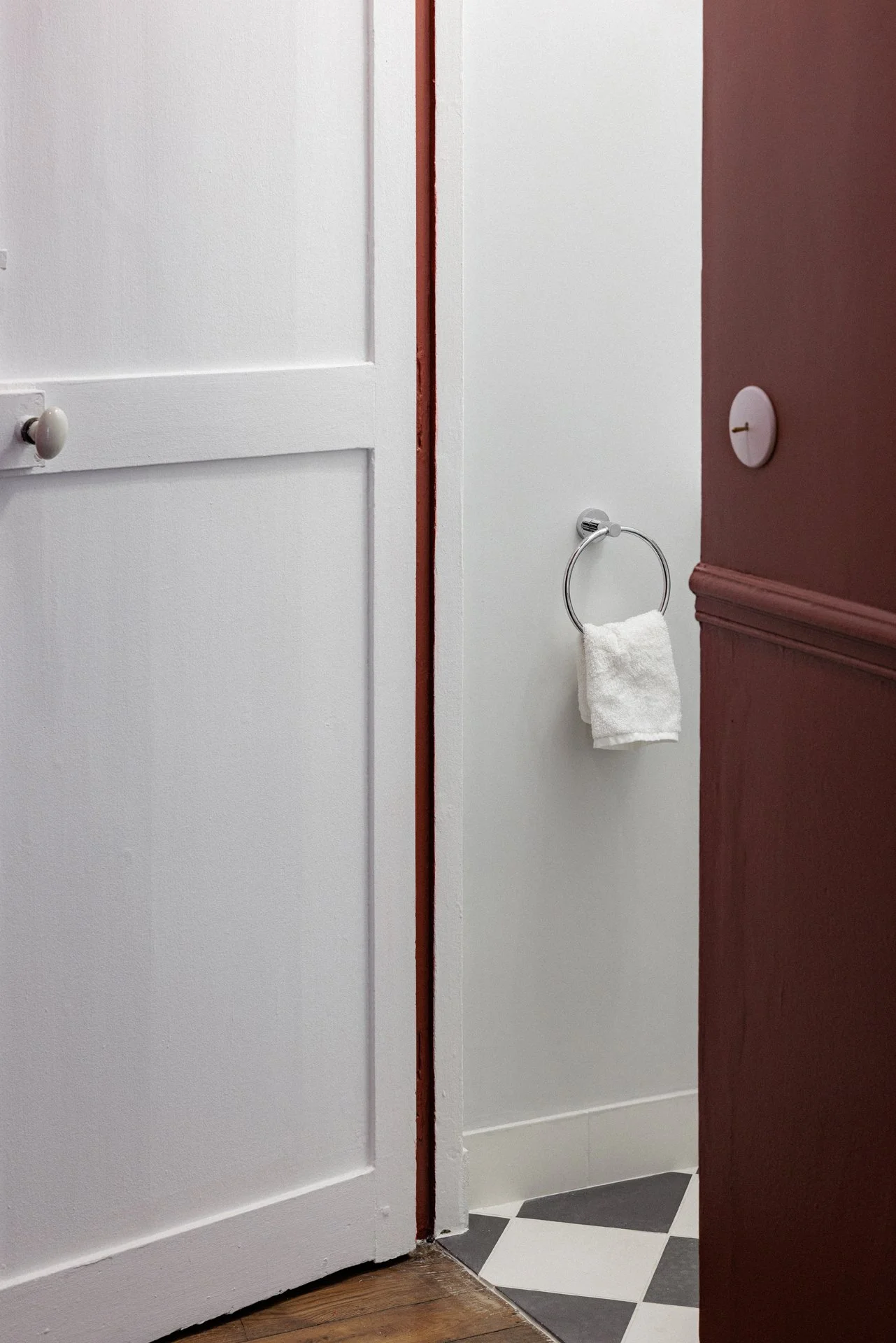 Enfilade de portes vers une salle de bain au sol en damier, anneau porte-serviette chromé et boiseries à panneaux, sur fond de mur bordeaux profond.