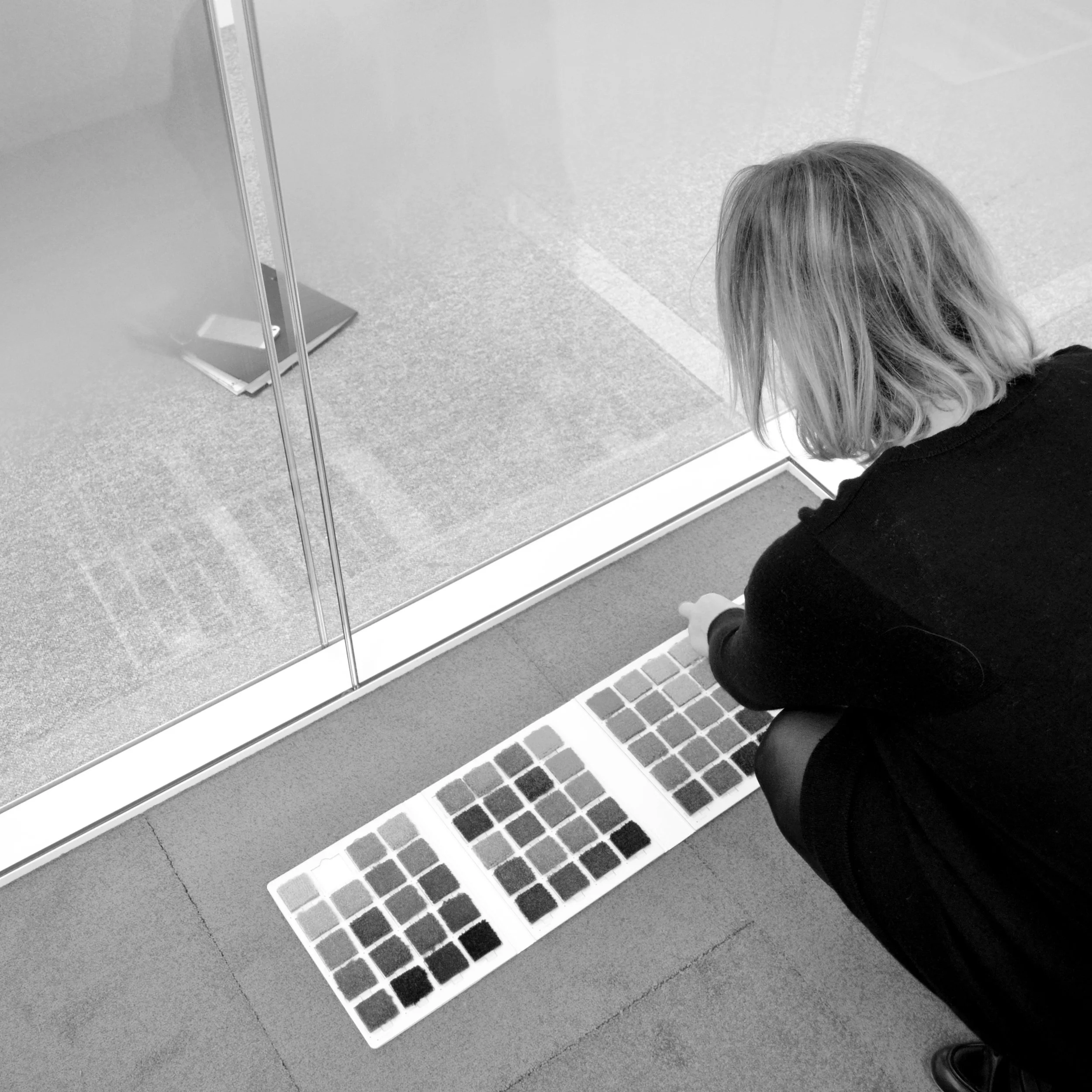 Photo en noir et blanc de Bea Bühler agenouillée, comparant des nuanciers de moquette dans un bureau vitré, sélection des matériaux pour l'aménagement des bureaux OCDE Boulogne