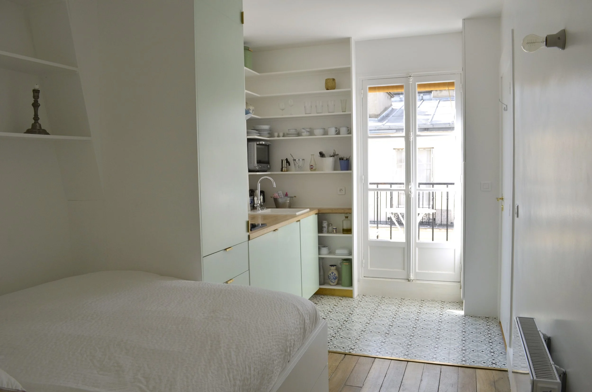 Vue d'ensemble du studio de 13 m2 : lit, kitchenette avec étagères ouvertes et porte-fenêtre donnant sur le balcon