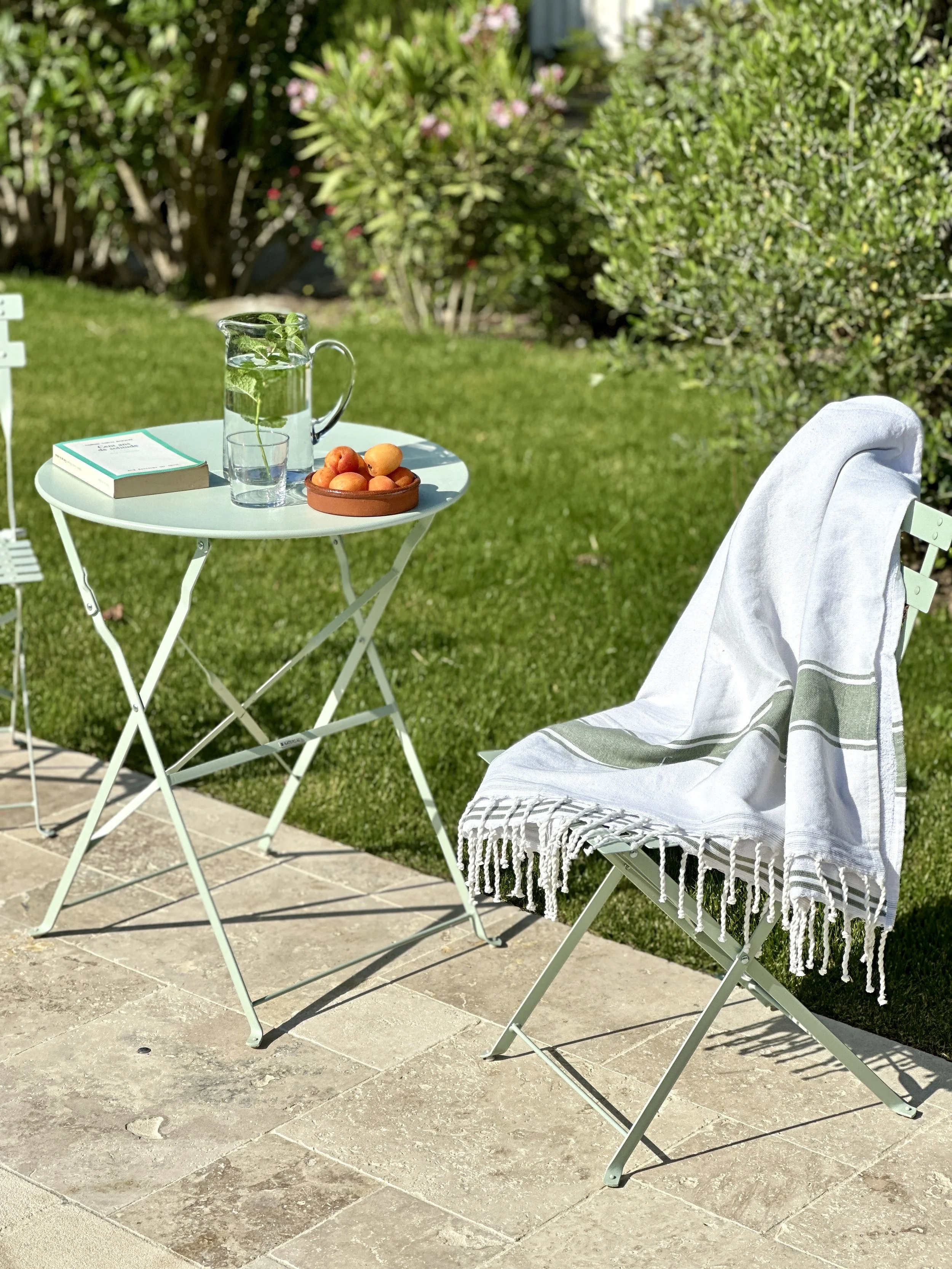 Table de jardin bistrot vert menthe avec pichet d'eau, abricots et livre, serviette de plage à franges sur chaise pliante, jardin ensoleillé