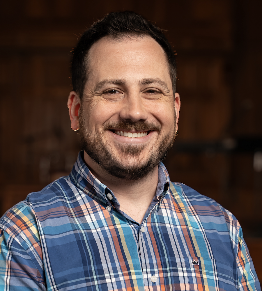 A smiling man in a colorful plaid shirt standing indoors with a wooden background.