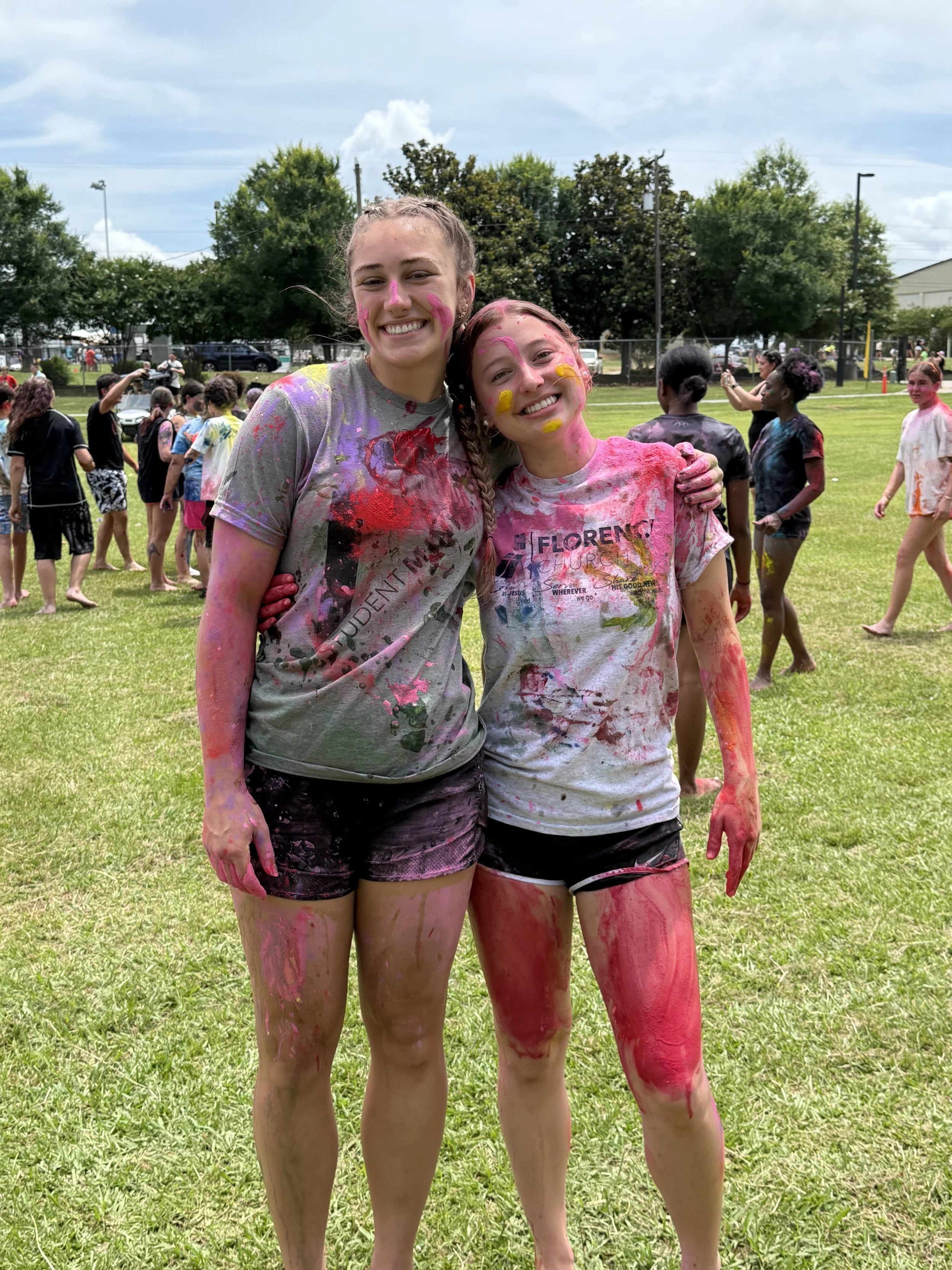 Two girls smiling and covered in multicolored powder at an outdoor event, with other participants in the background.