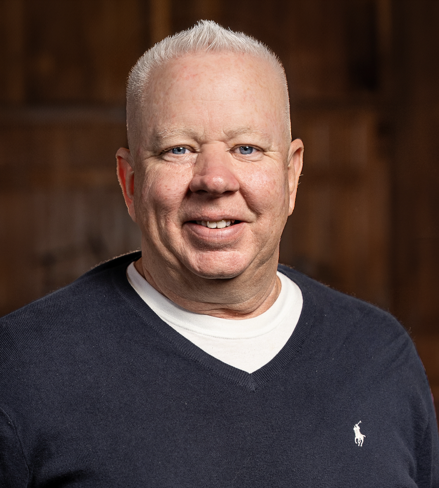 Portrait of a smiling middle-aged man with short white hair, wearing a navy blue sweater with a Ralph Lauren logo, and a white t-shirt underneath, in a warmly lit indoor setting.