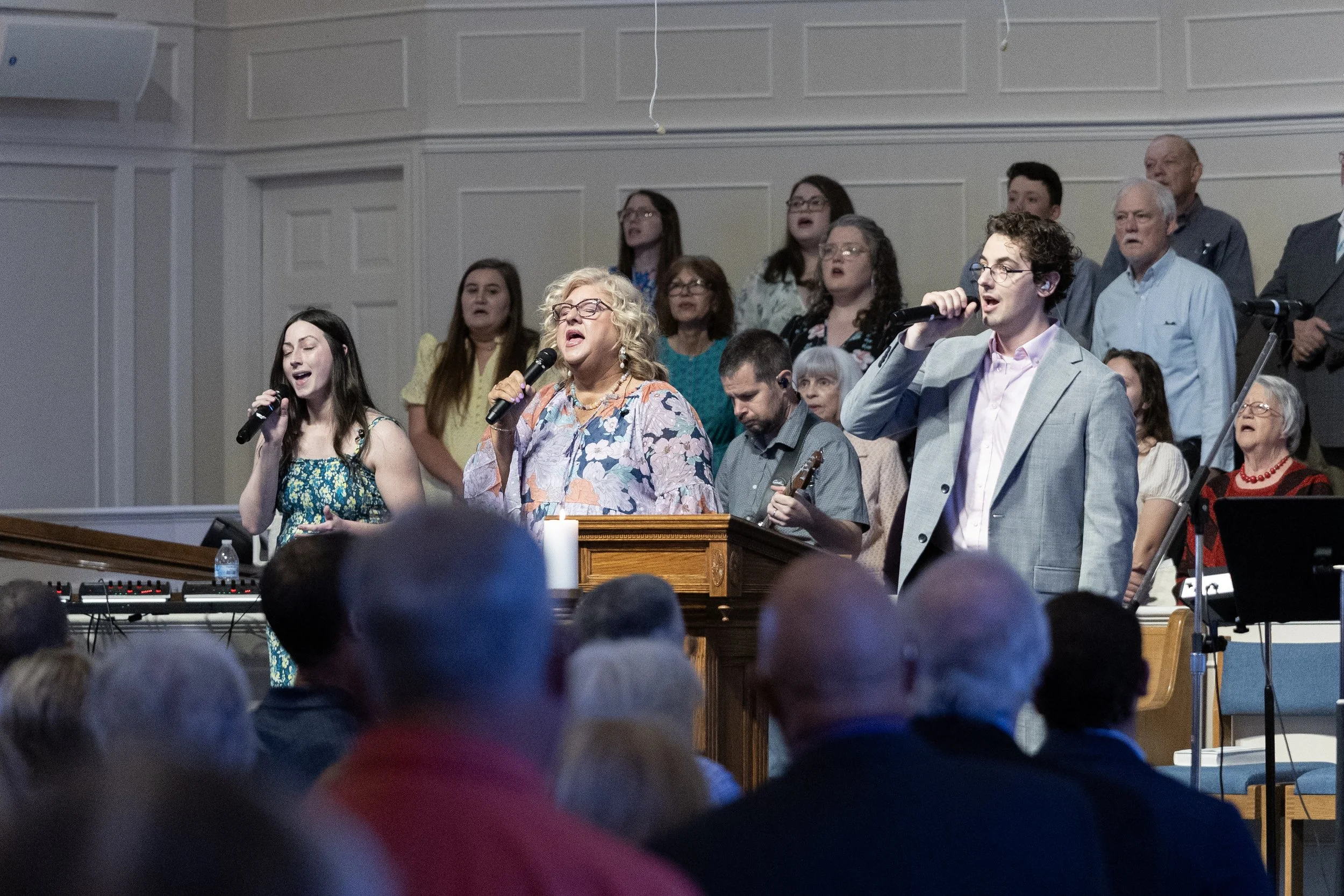 A group of people in a church choir singing with some holding microphones, standing behind a pulpit, with an audience visible in the foreground.