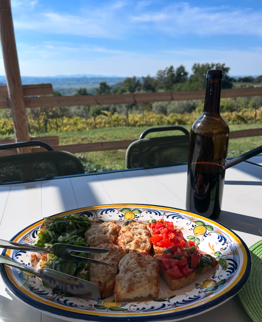 Platter of three varieties of bruschetta beside a glass of red wine, overlooking the Tuscan countryside.