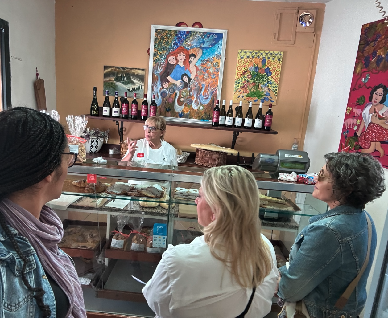 Three women speaking with a baker behind a glass display of breads and pastries in a small Italian bakery.