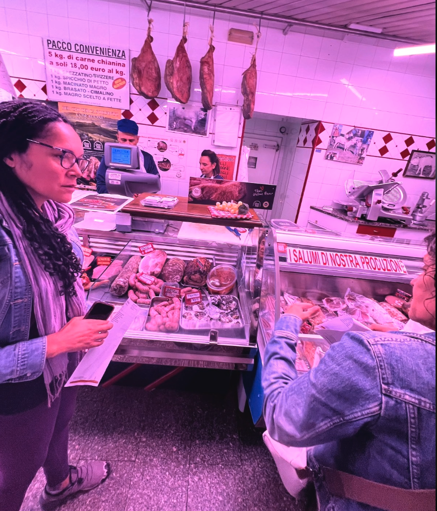 Customers shopping at a Tuscan butcher counter displaying sausages, cured meats, and fresh cuts of meat.