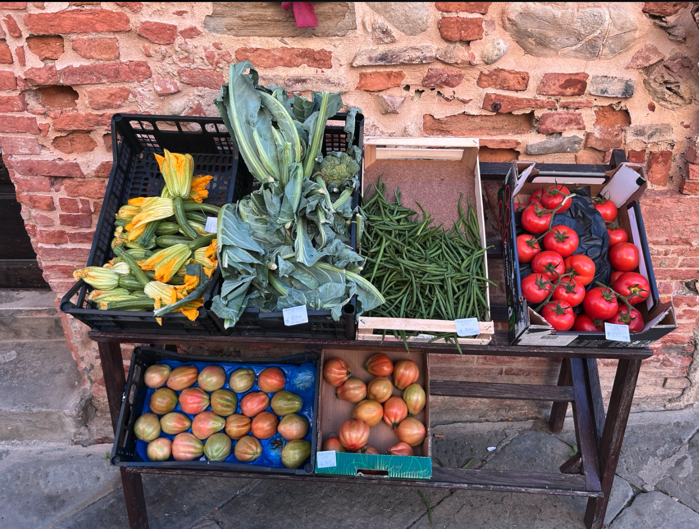 Wooden display of fresh tomatoes, green beans, leafy vegetables, and yellow squash blossoms outside an Italian shop.