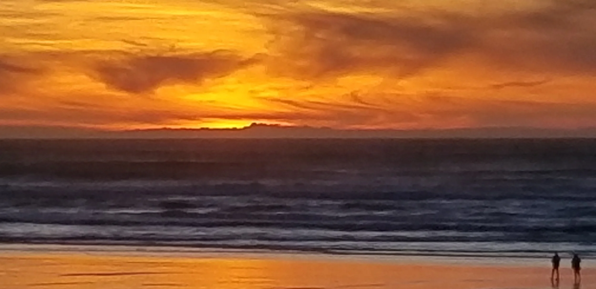 A sunset over the ocean with a cloudy sky, a few people walking on the beach in the foreground.