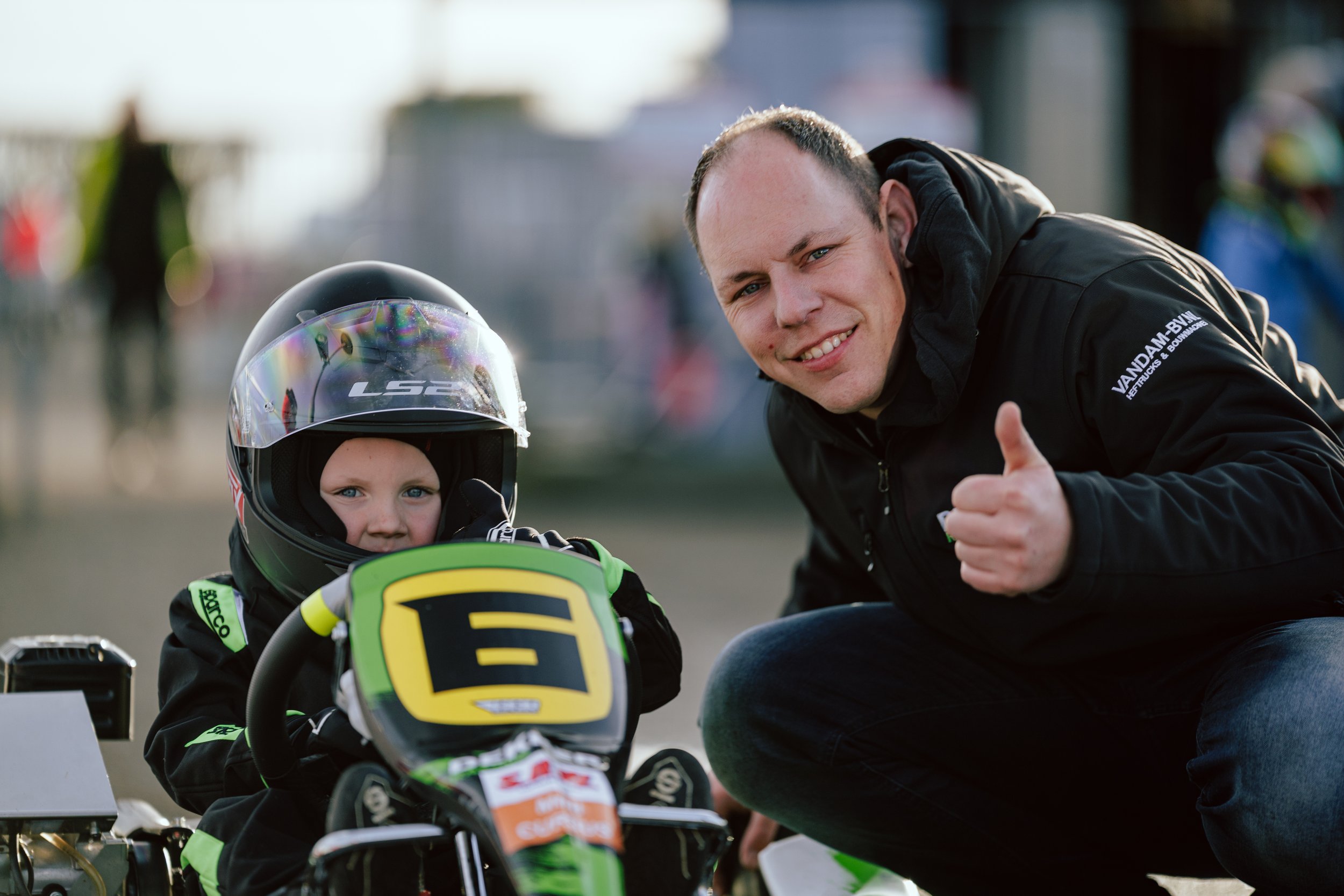 A young go-kart racer in a black racing suit with a helmet, sitting in a go-kart with the number 5, alongside an adult man crouching next to them, giving a thumbs-up gesture.