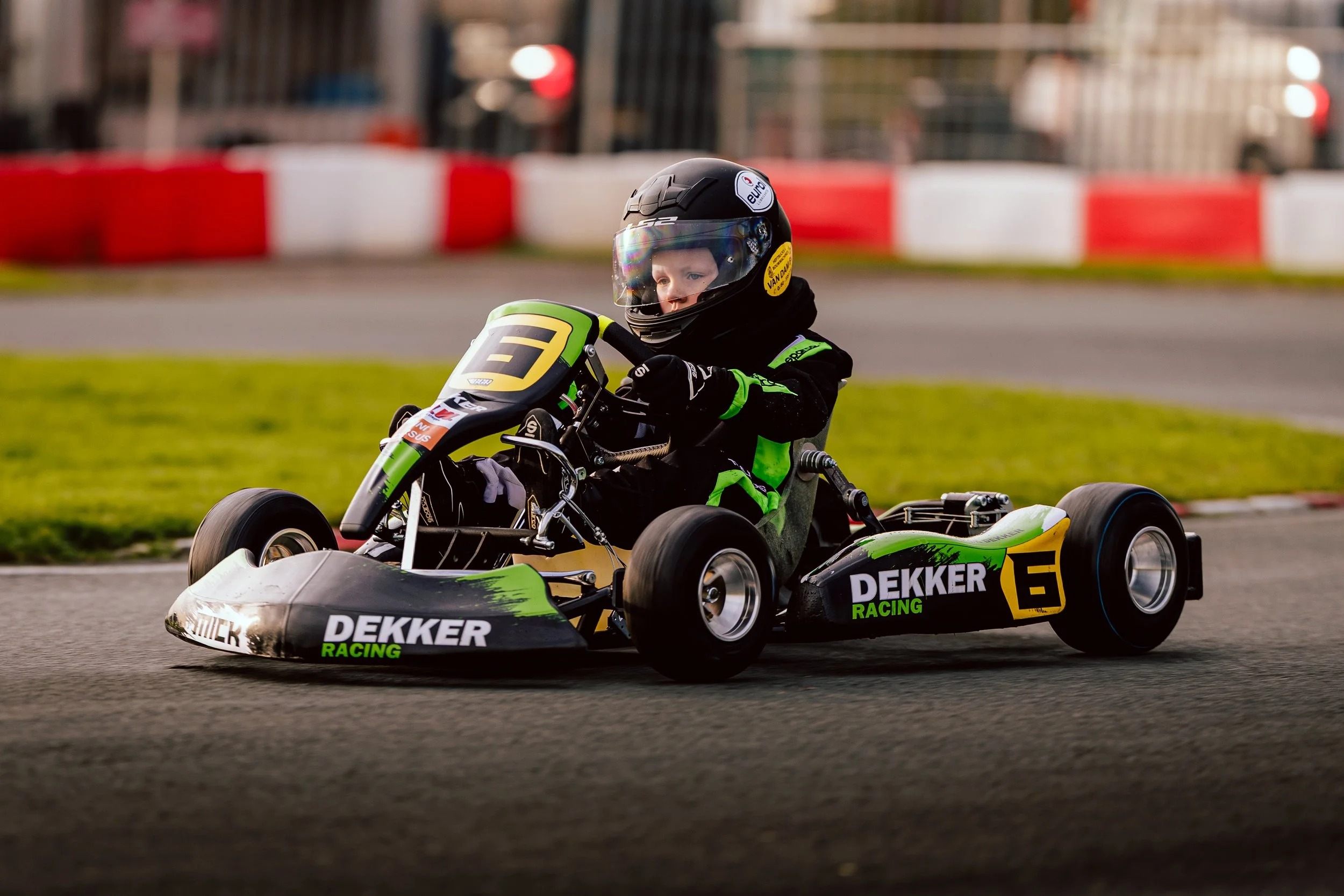 Young child racing a go-kart on a race track, wearing a black helmet and racing suit, with green and black go-kart labeled 'Dekker Racing' and the number 6.