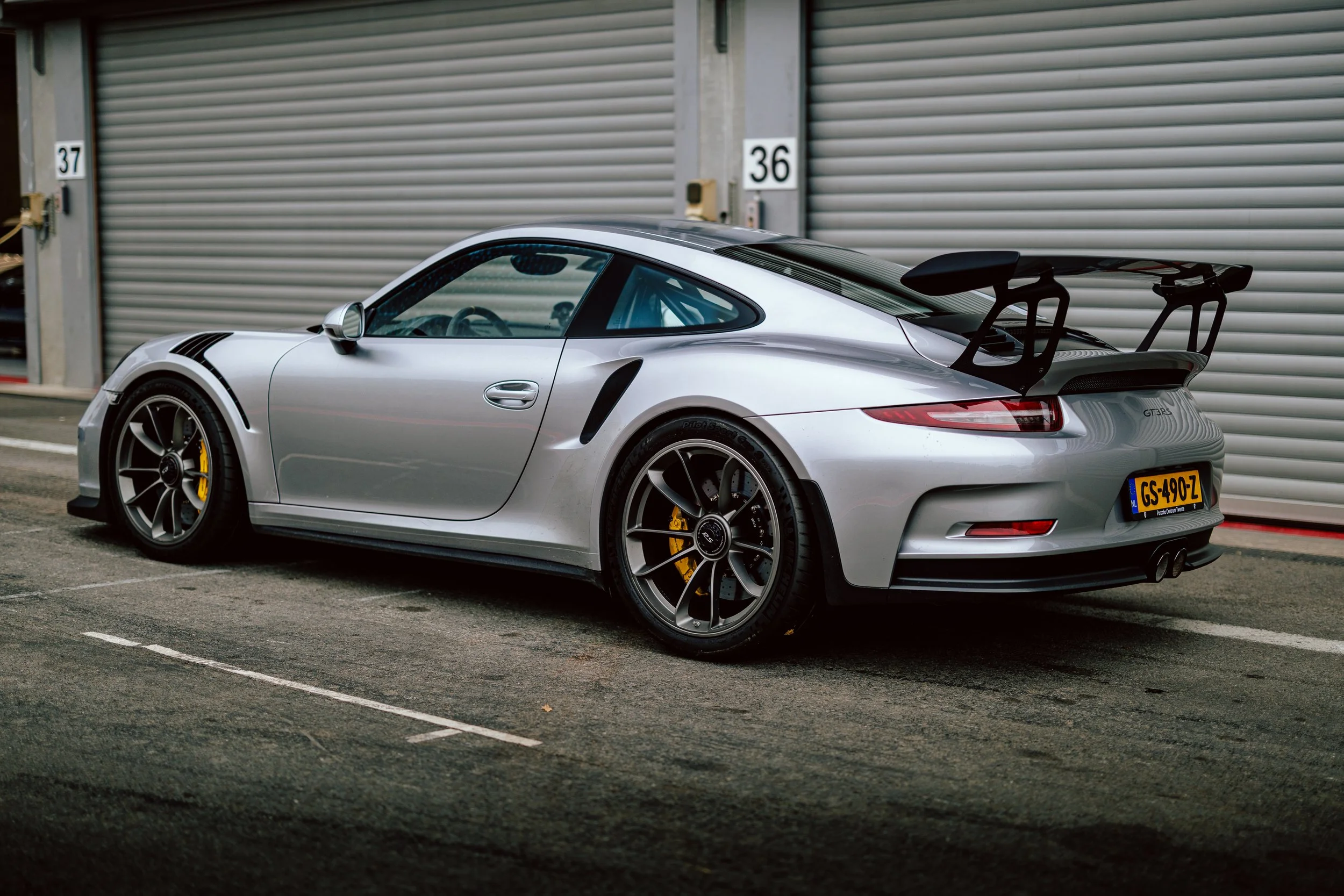 Silver sports car, Porsche 911 GT3 RS, parked outside a building with closed metal shutters, featuring a large rear wing and black wheels with yellow brake calipers.