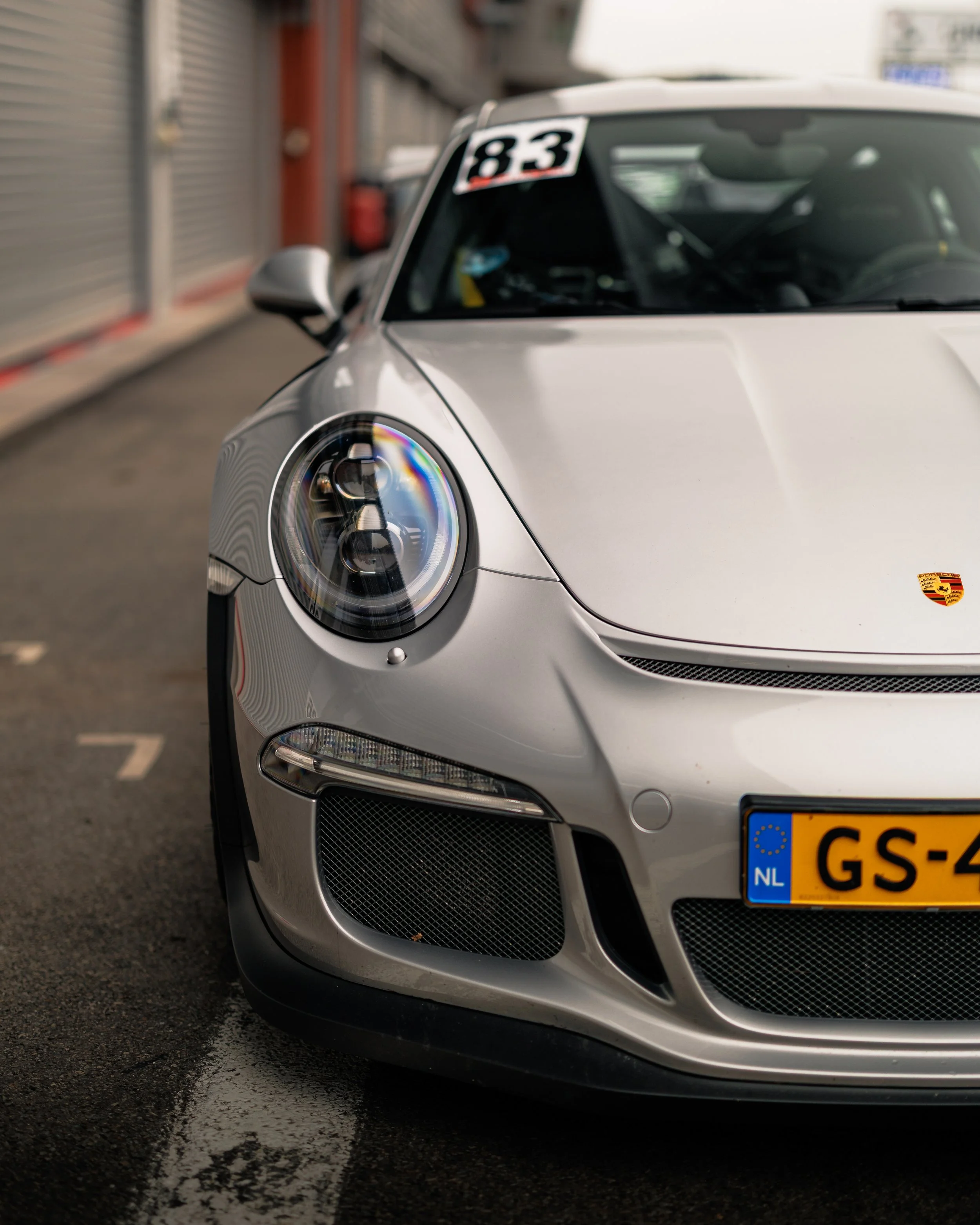 Close-up of front of silver Porsche 911 race car with number 83 on windshield, parked on racing track with empty garages in background.
