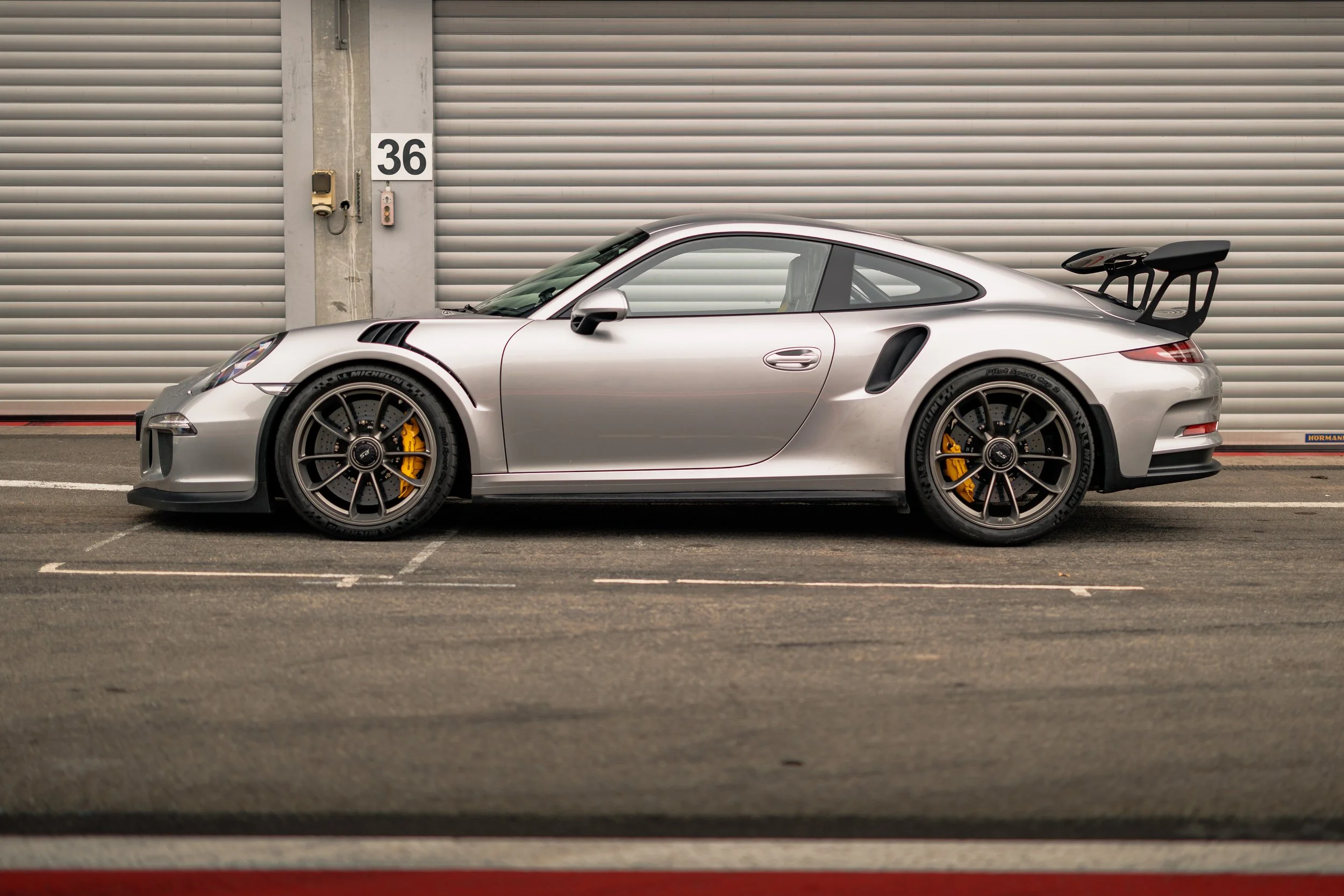 Silver Porsche 911 GT3 sports car with rear wing parked in a garage near a large roll-up door.