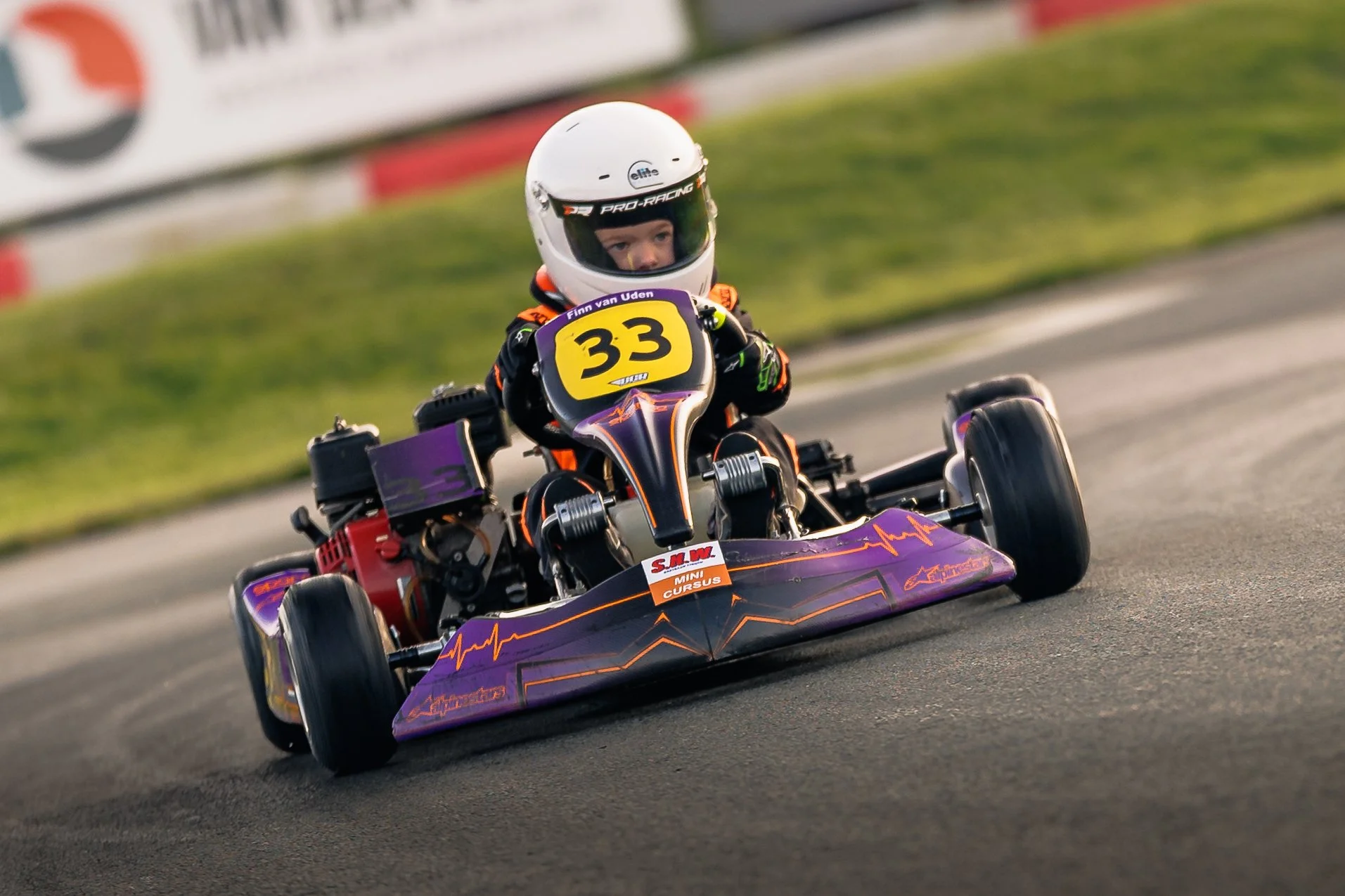 Young boy racing in a purple and black go-kart with the number 33, wearing a white helmet and racing suit, on an asphalt track.