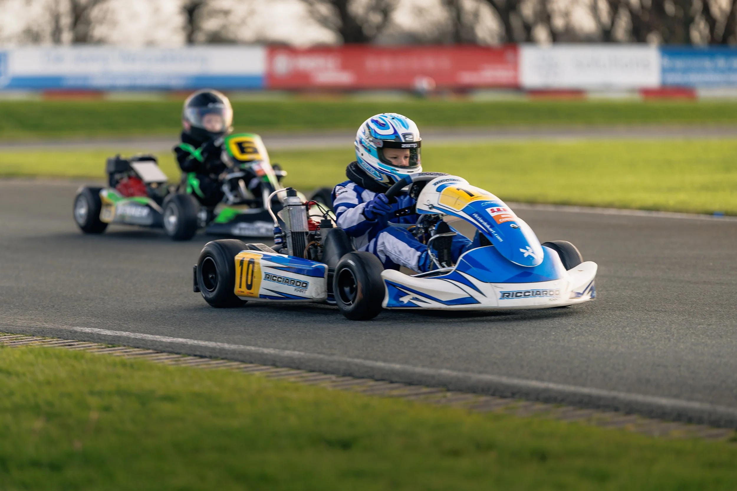 Two children racing go-karts on a track, wearing helmets and racing suits, with a grassy infield and track barriers in the background.
