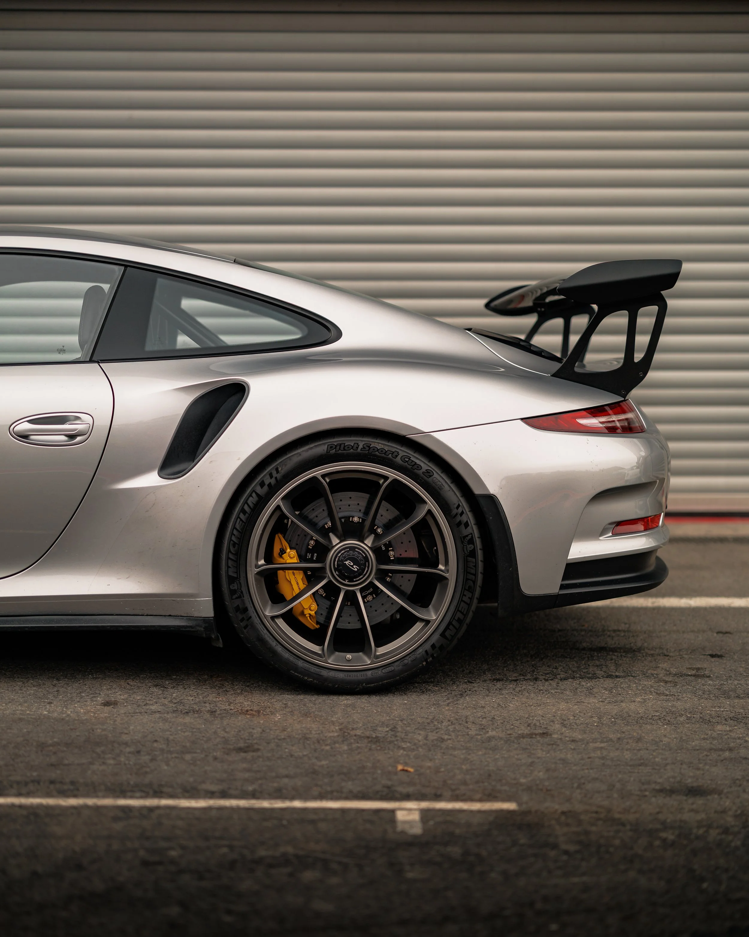 Silver sports car with black rear wing, yellow brake calipers, parked on asphalt in front of a metallic garage door.
