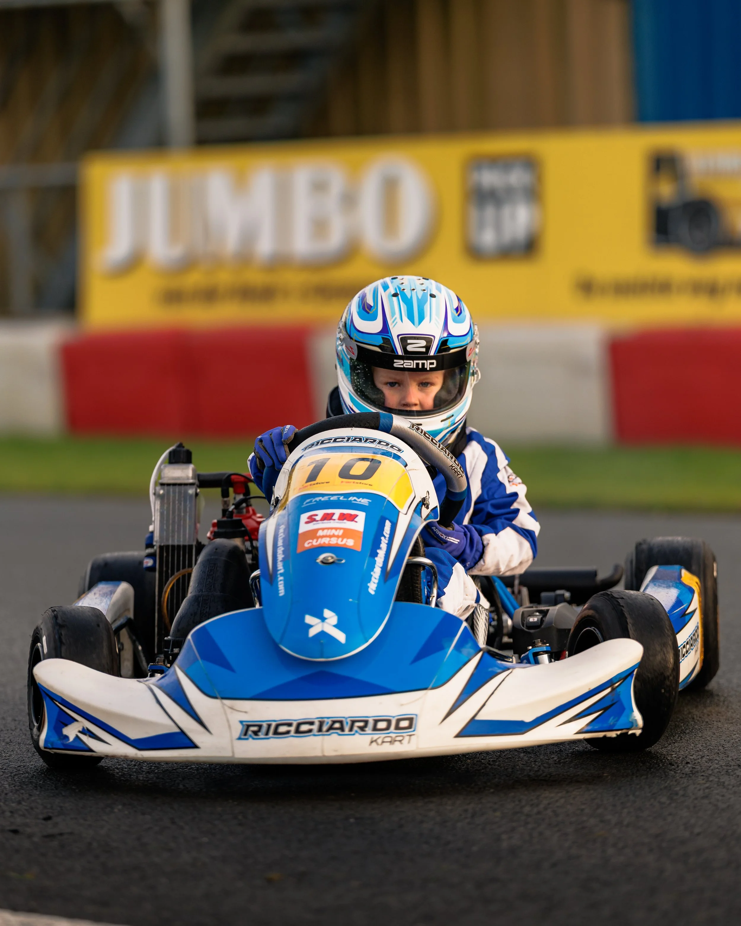 A young child racing a blue and white go-kart on a track, wearing a helmet and racing suit, with a blurred yellow and red advertisement in the background.