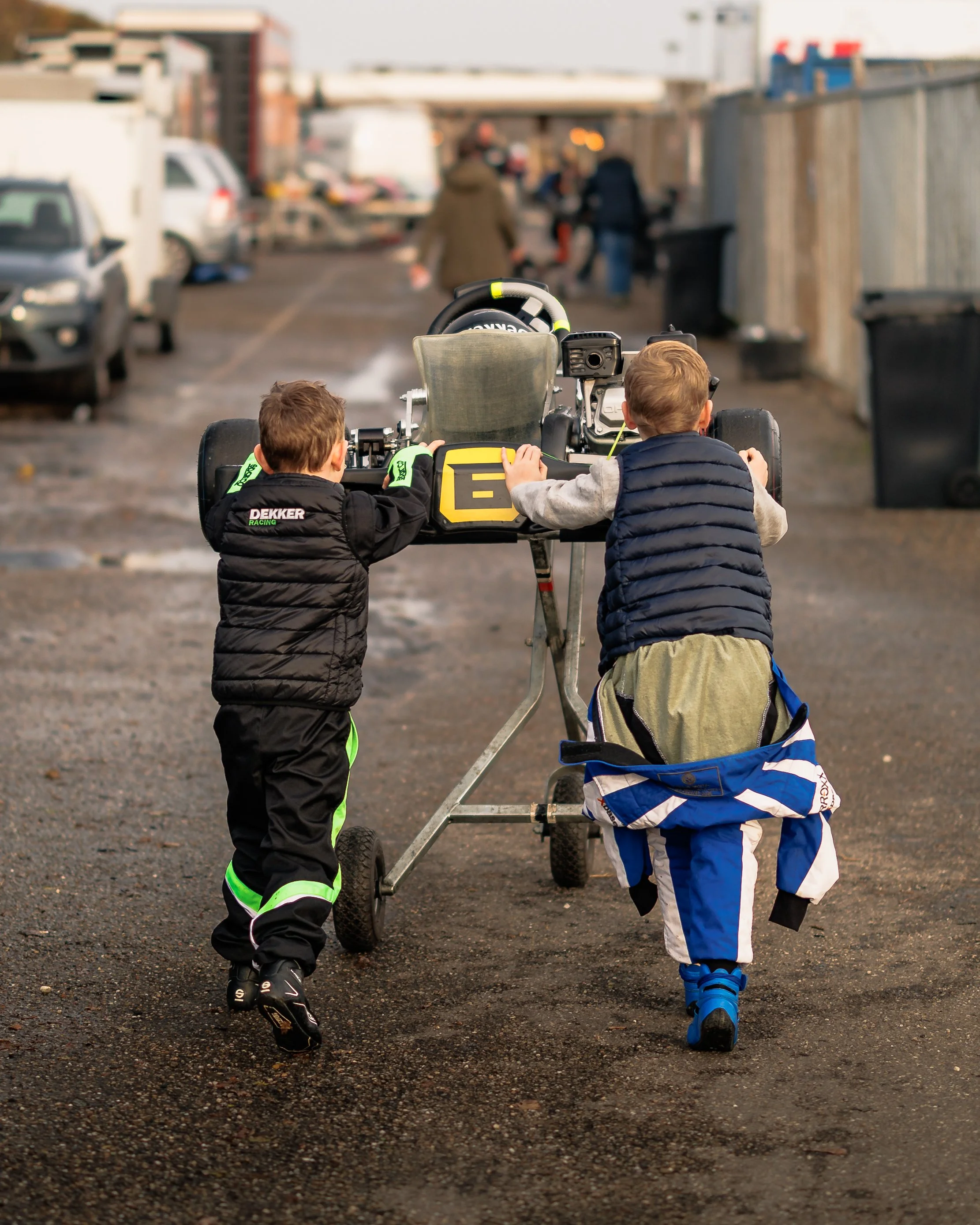 Two young boys pushing a racing go-kart down an alleyway. One boy is wearing a black racing suit with green accents, the other is wearing a blue and white racing suit with a gray vest. The alley has parked cars and people in the background.