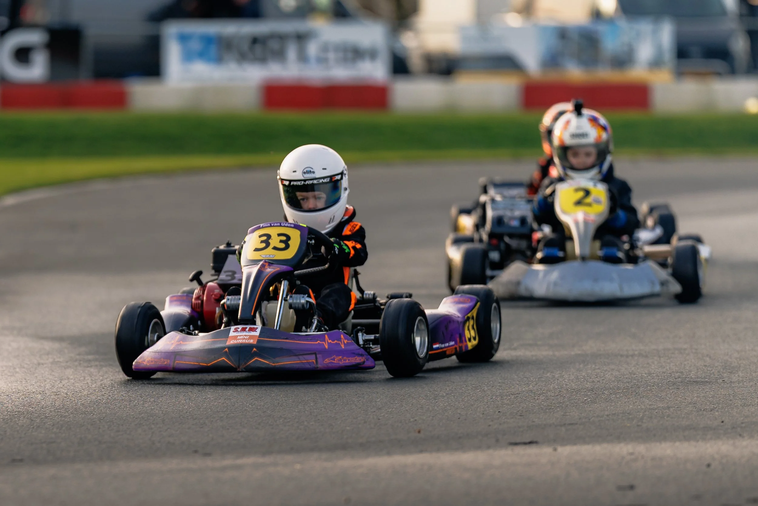 Three young go-kart racers in helmets and racing suits on a paved track, with the lead driver in kart number 33 closely followed by kart number 2 during a race.