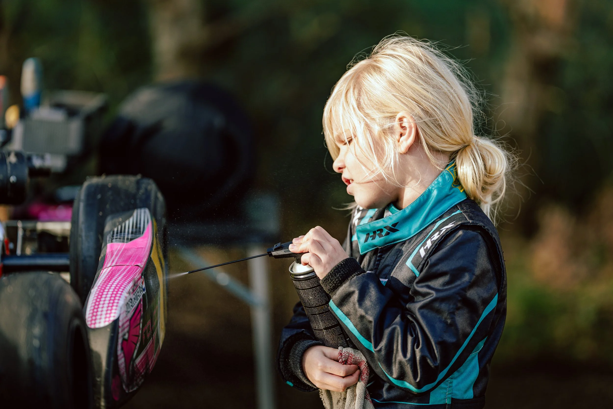 A young girl with blonde hair in a ponytail, wearing a black and teal racing suit, is spray cleaning her dirt bike outdoors in a wooded area.