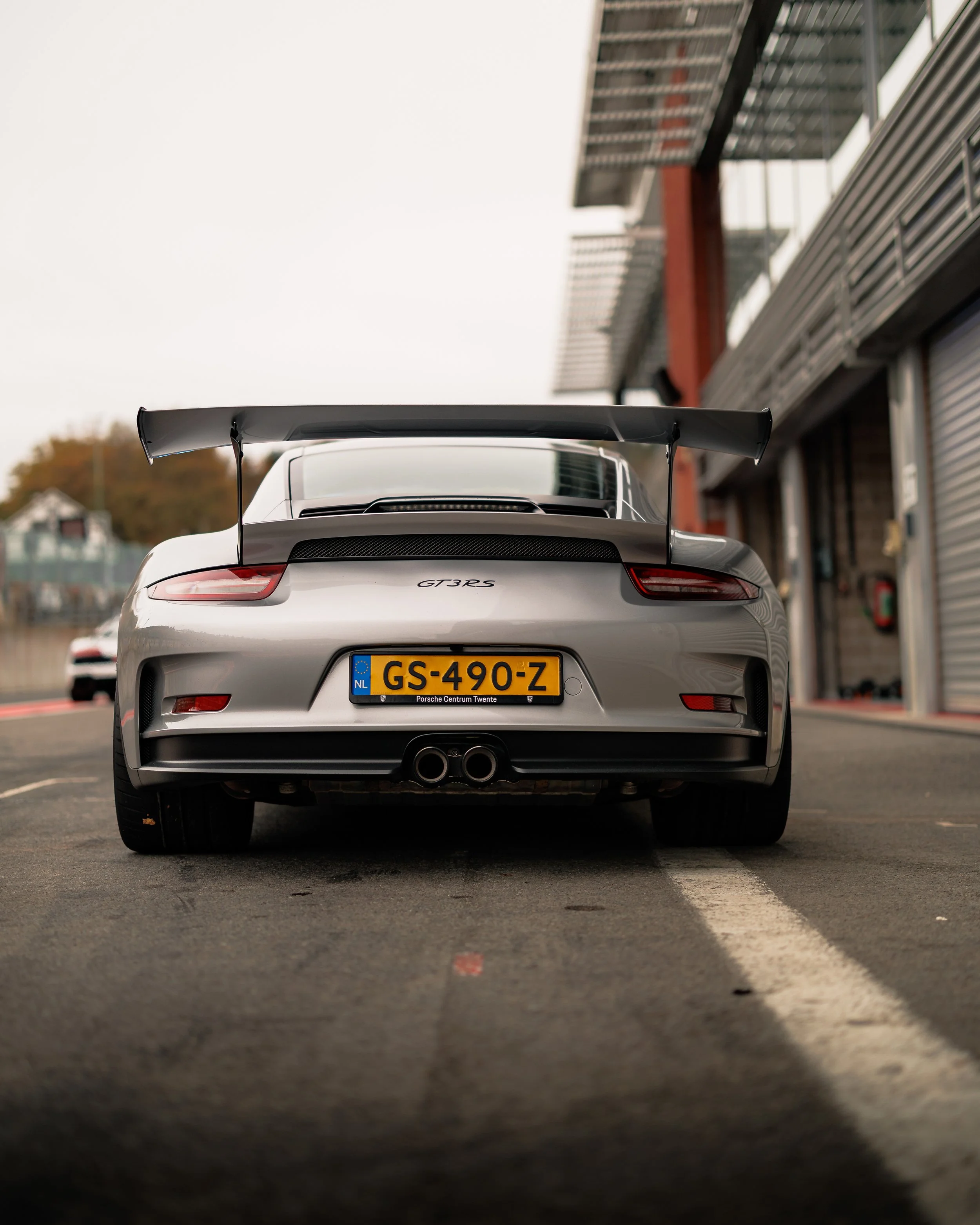 Rear view of a silver Porsche 911 GT3 RS parked on a race track with a large rear wing, dual exhaust pipes, and a yellow Dutch license plate.