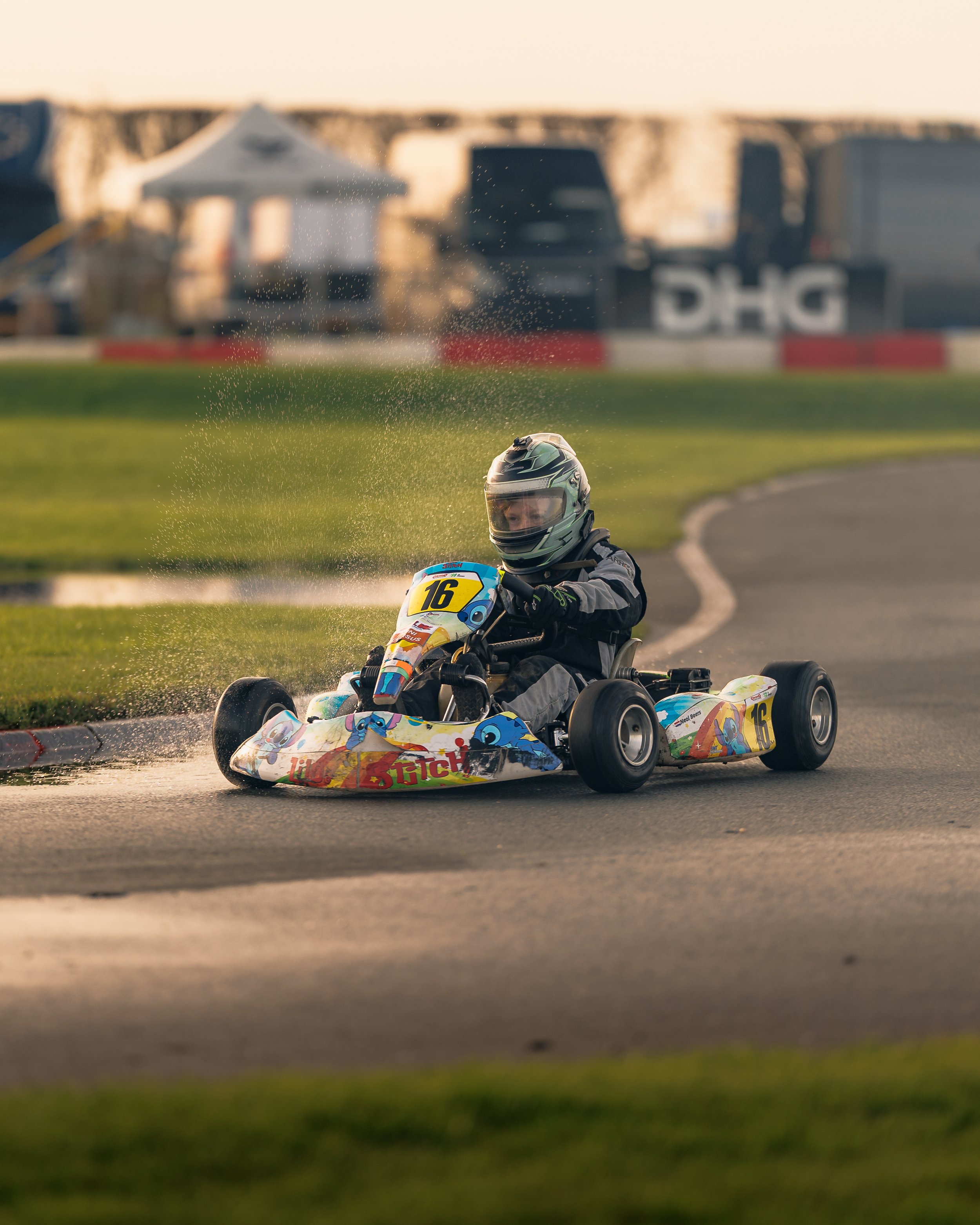 A person in a helmet driving a go-kart on a racetrack, splashing water on the track during a race.