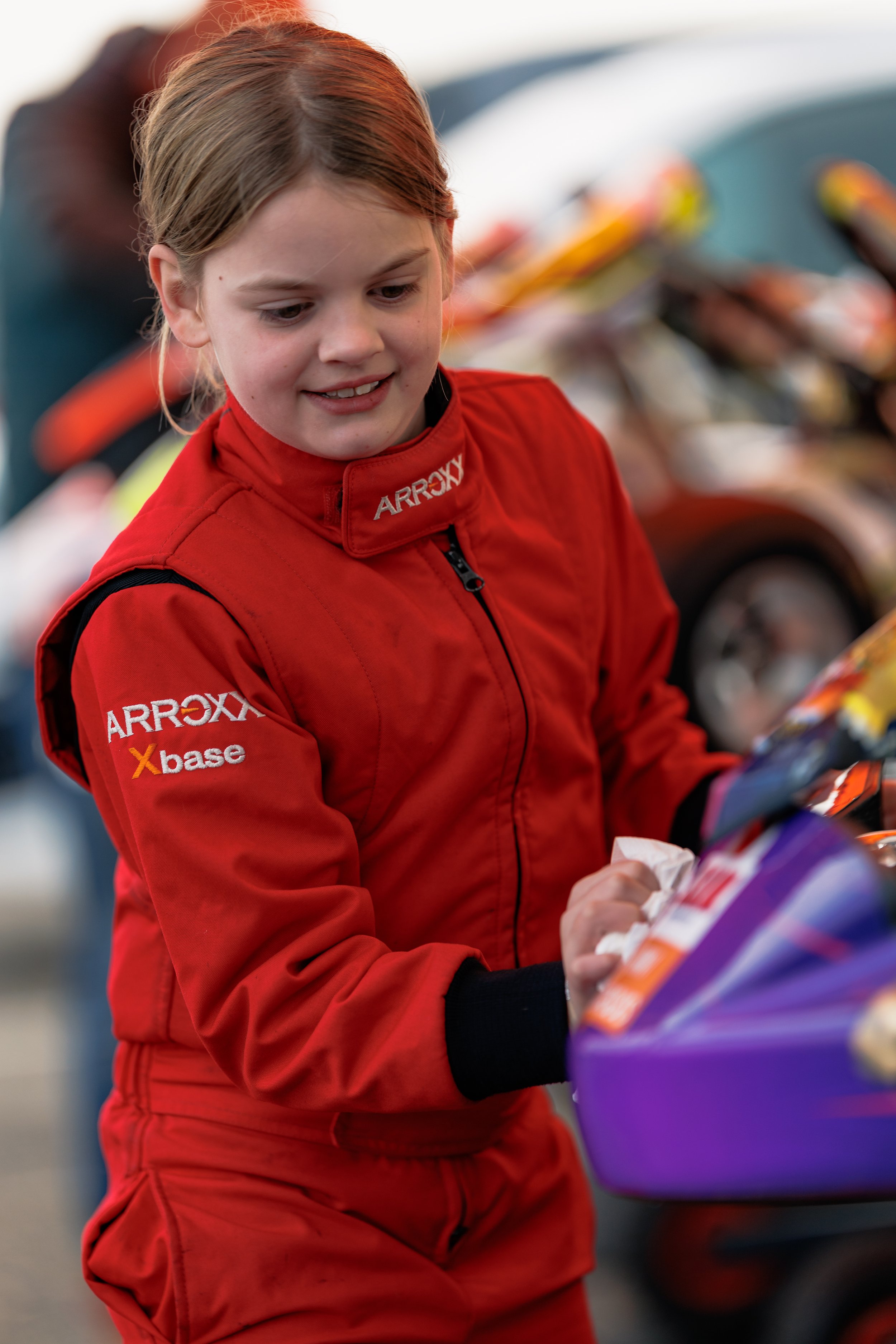 A young girl in a red racing suit is smiling and selecting snacks from a display, likely at a racing event.