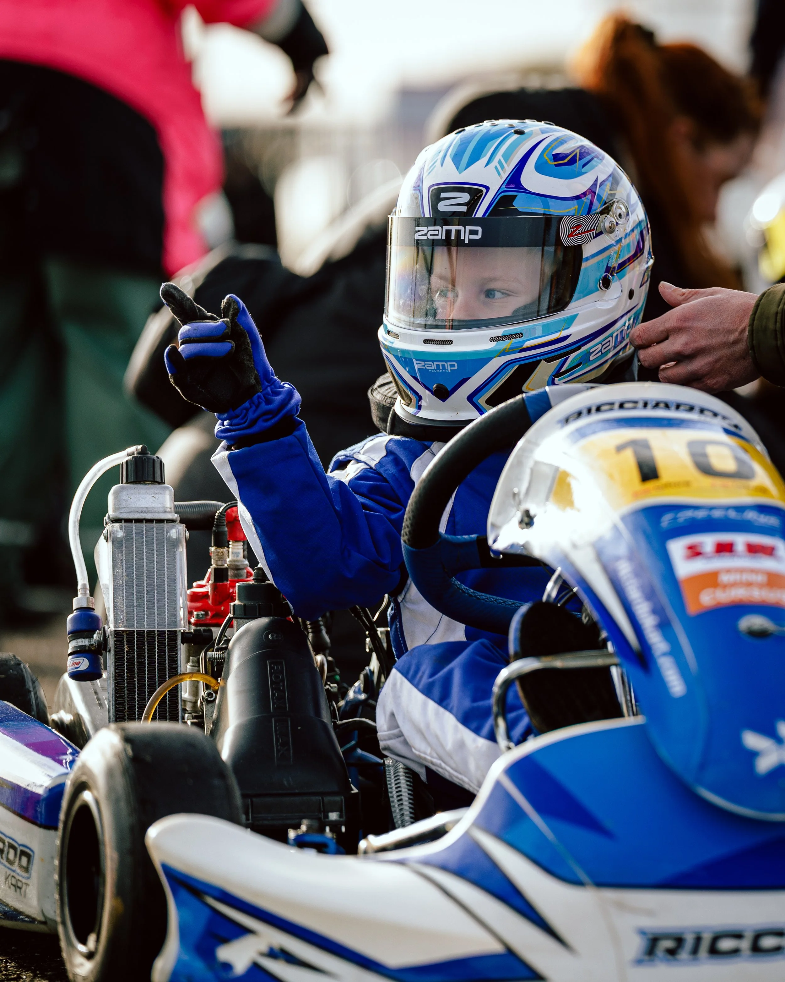 A young race car driver sitting in a blue and white go-kart, wearing a blue racing suit and helmet, giving a thumbs-up while being assisted by someone.