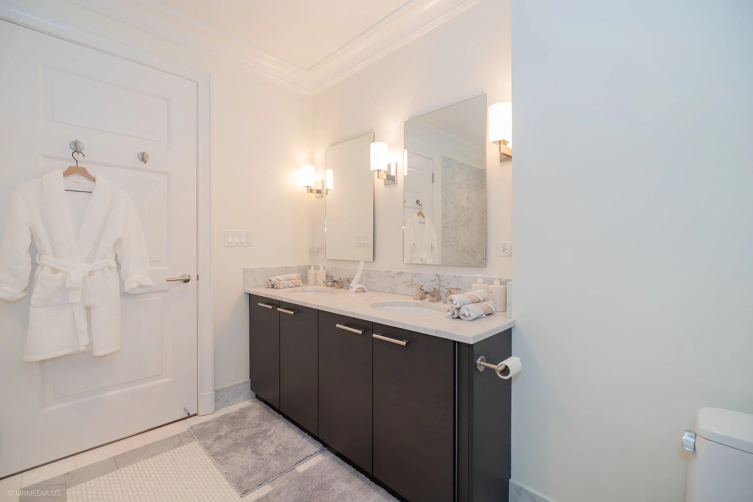 Modern bathroom with double sink vanity, marble countertop, mirrors, wall sconces, bathrobes, towels, and tile flooring.