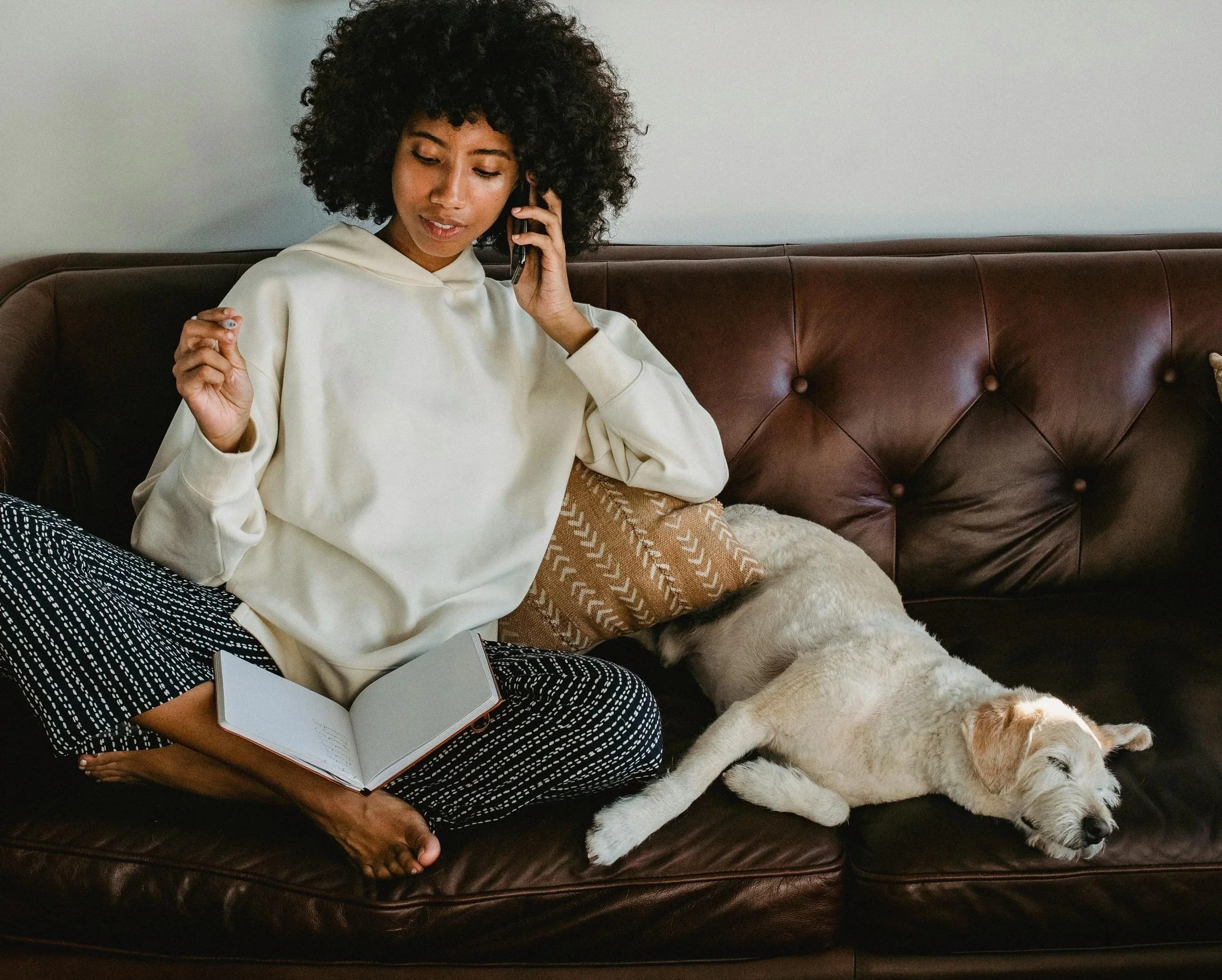 A woman with curly hair sitting on a leather couch, talking on a phone, with a notebook in her lap and a dog lying nearby.
