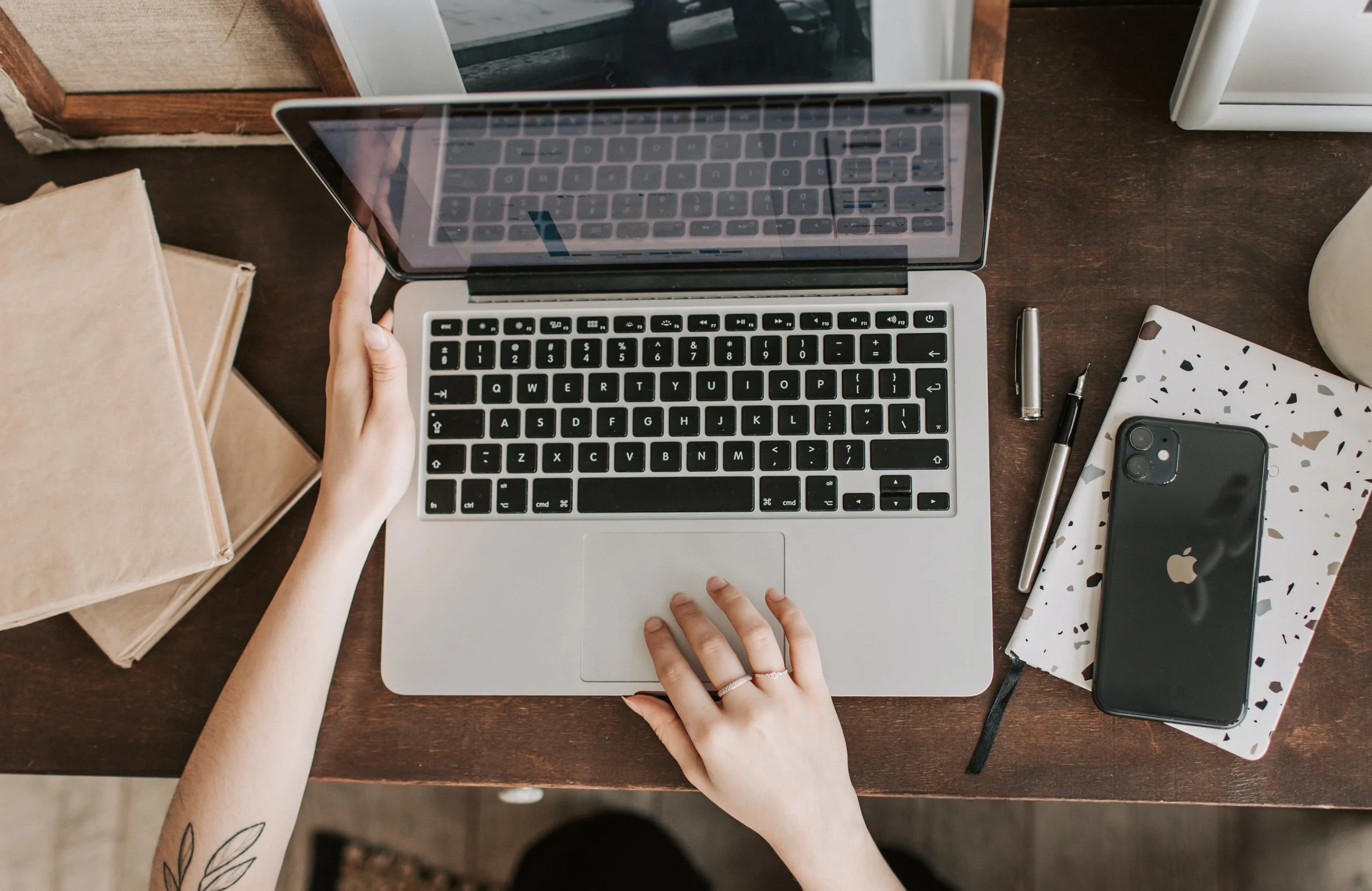 Top-down view of a person with tattooed arms using a silver laptop at a wooden desk, with a smartphone, notebook, pens, and decorative items nearby.