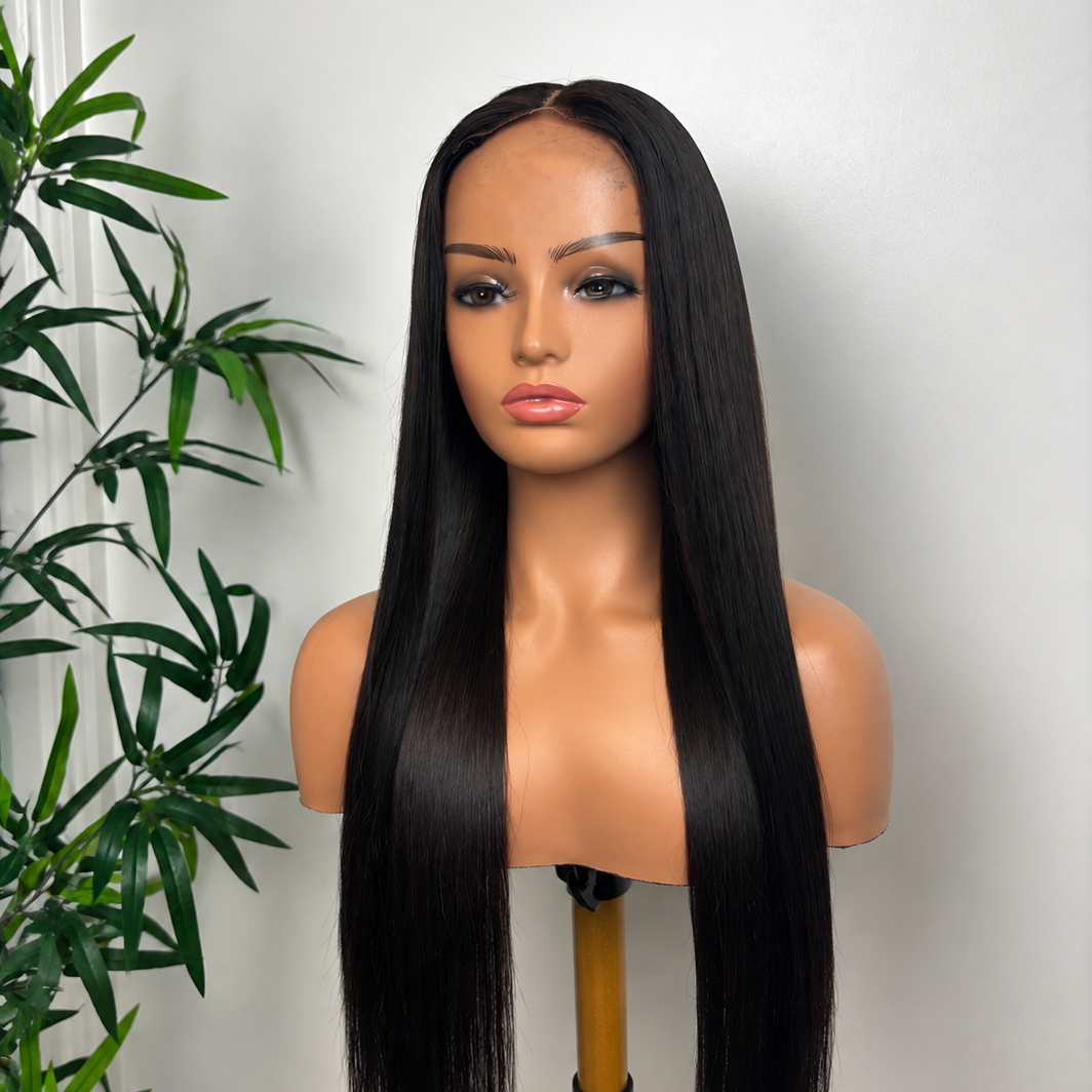 A mannequin with long, straight black hair, makeup, and a neutral expression, positioned beside a green plant against a plain wall.