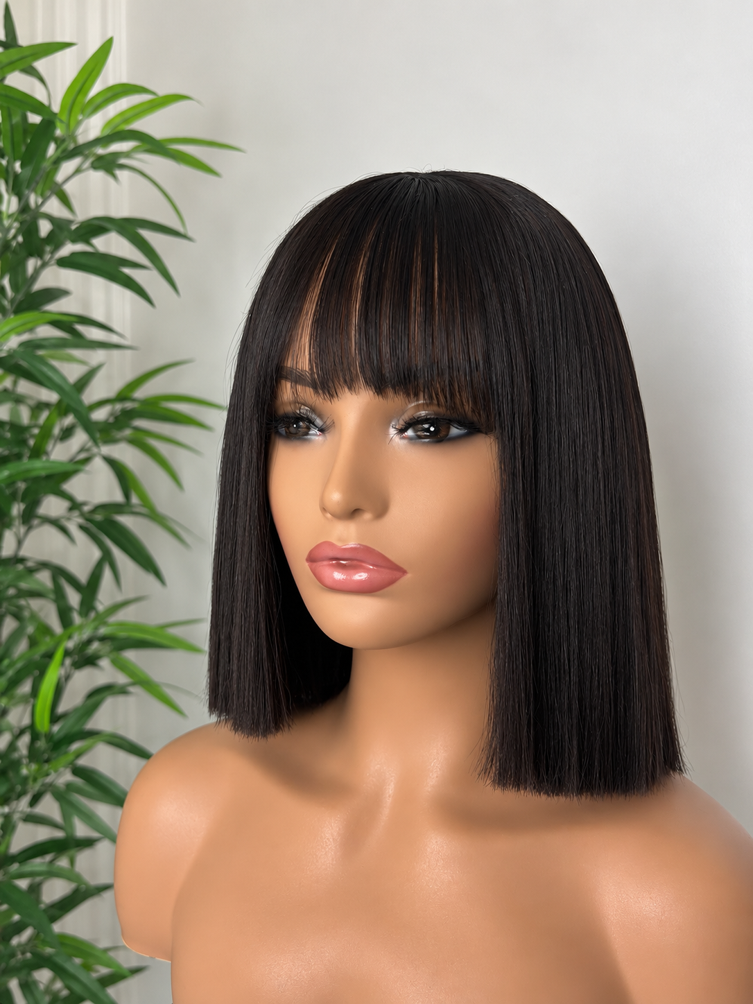 A realistic female mannequin with straight, shoulder-length dark brown hair with bangs, positioned in front of a white wall and a green leafy plant.