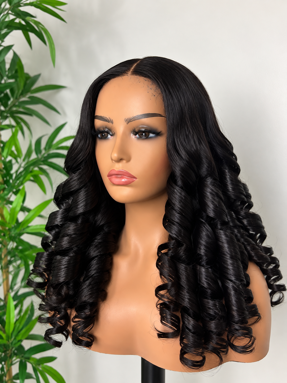 Mannequin with long, black, curly hair posing in front of a white background and green plant.