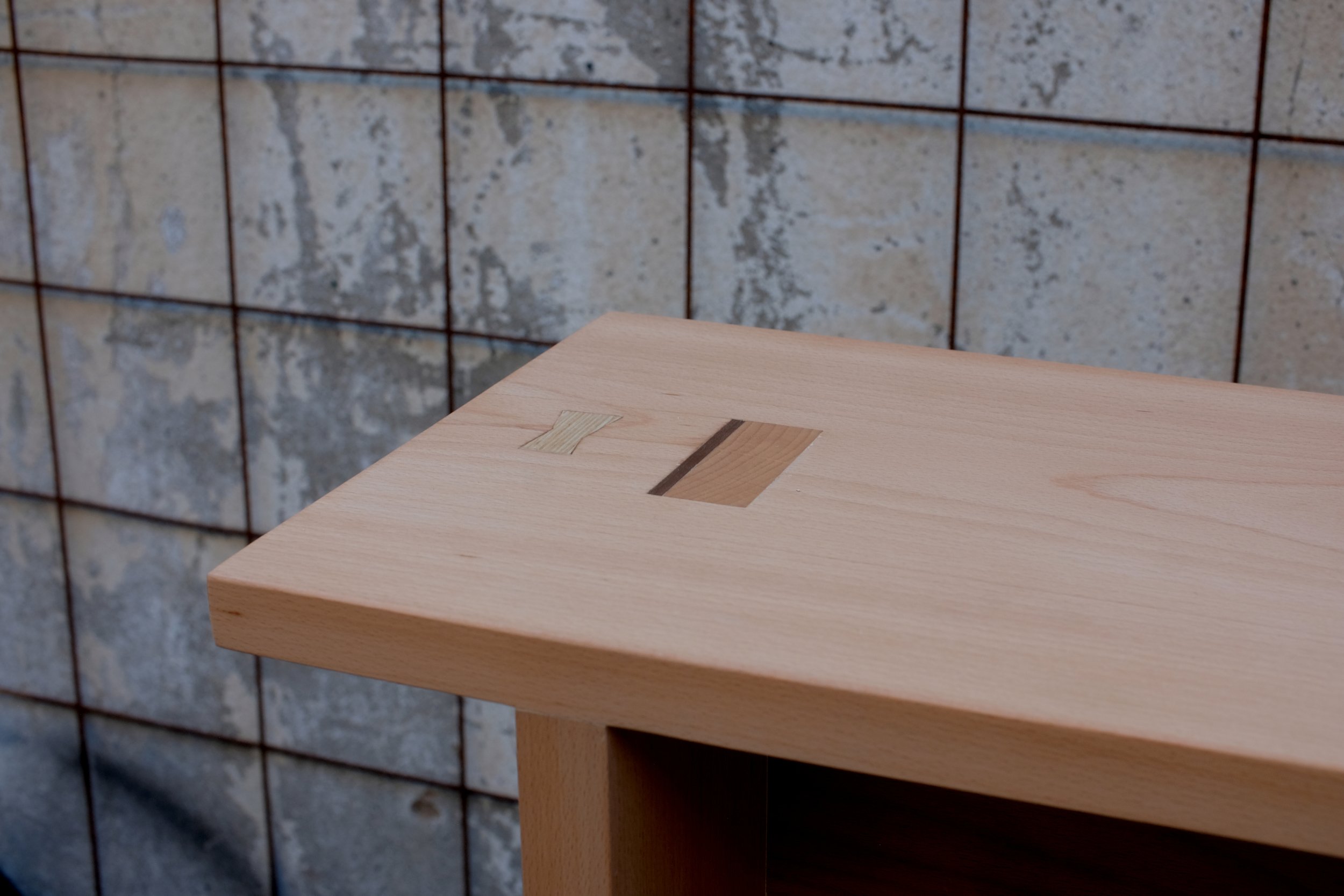 Close-up of a light wood bench top with a composite through tenon joint on the top surface, made of light and dark wood. The background features a textured stone wall with a metallic grid.